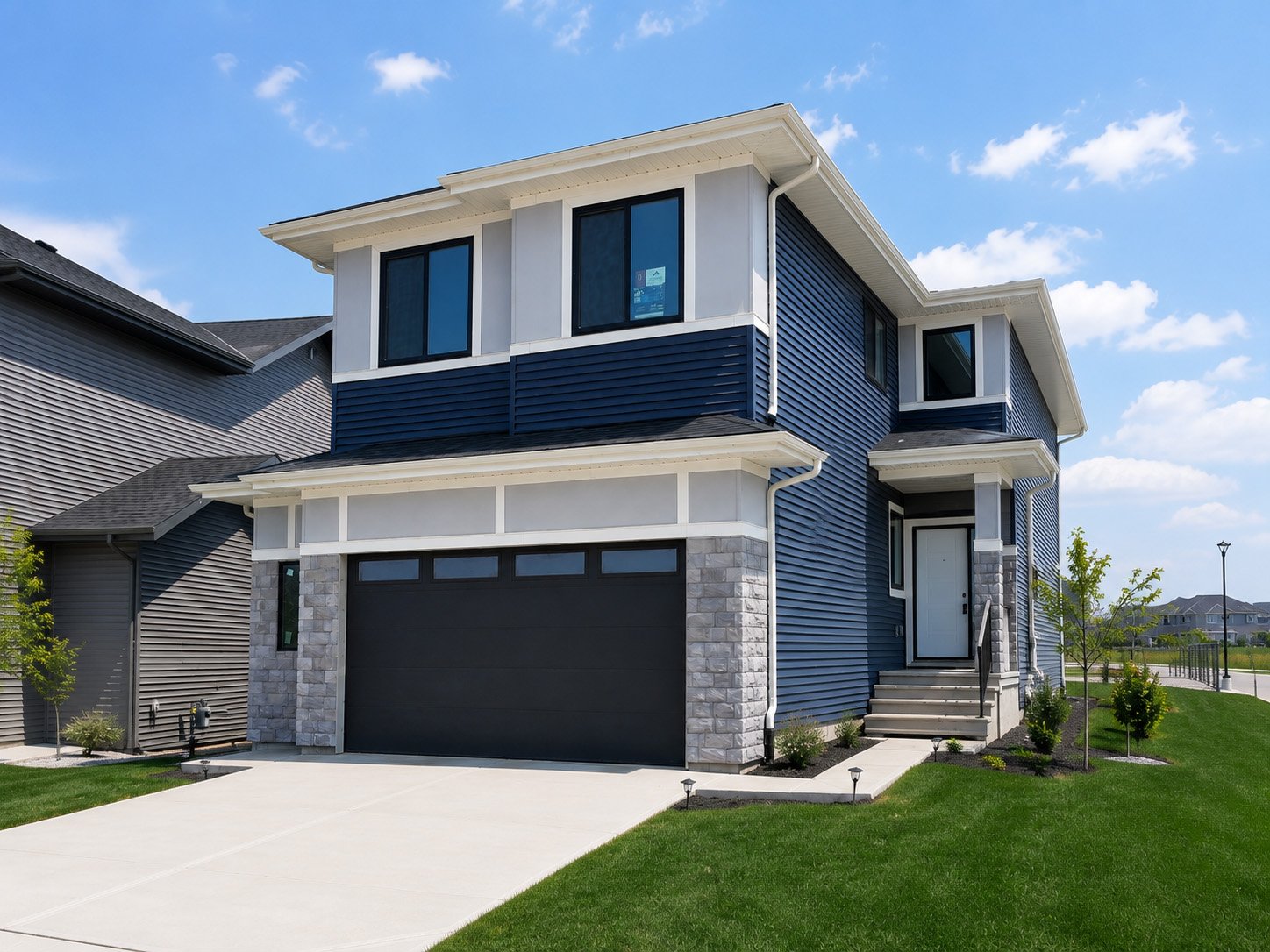 Modern two-story house with blue siding, gray stone accents, black garage door, and small front porch with steps, surrounded by a well-maintained lawn and young trees under a blue sky.