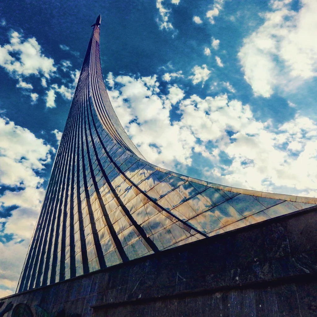 A tall modern metal monument commemorating space exploration with the depiction of a rocket taking off, the flames of the rocket serve as the support for the rocket sculpture atop. All set against a partly cloudy sky.