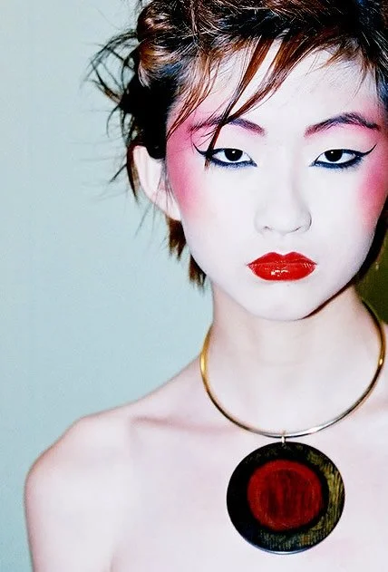 A woman with short hair styled in an edgy cut, wearing bold makeup including red lipstick and dramatic eye makeup. She has pink blush on her cheeks, a gold necklace with a large circular pendant, and is posing against a plain background.