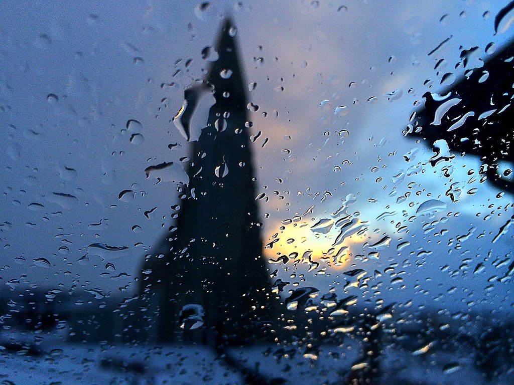 Rain-covered window with droplets of water, with a blurry silhouette of a tower or building and a cloudy sky in the background.