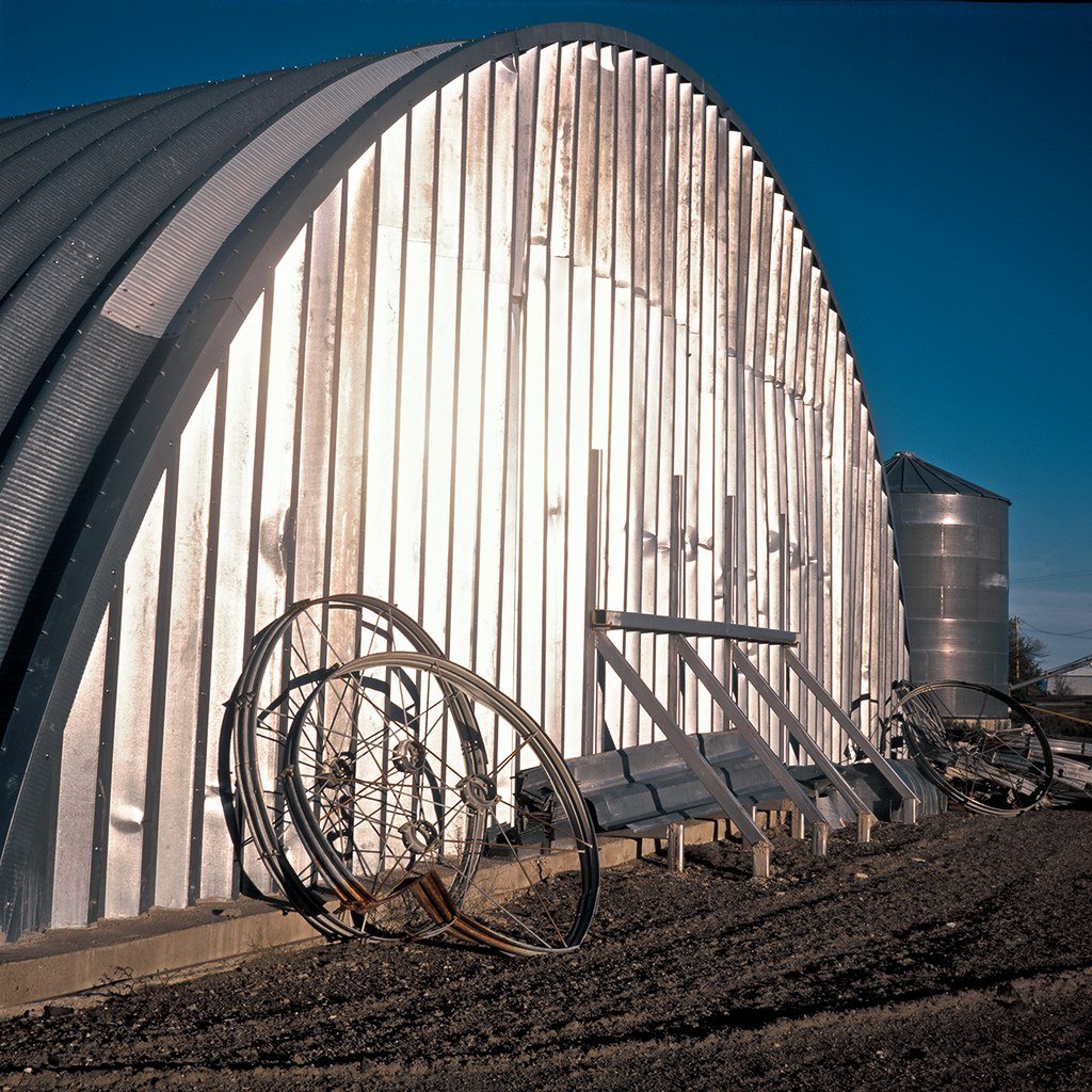 Metalized barn with curved roof and vertical panels, with old bicycle wheels leaning against it, set outdoors on dirt ground, under a clear blue sky.