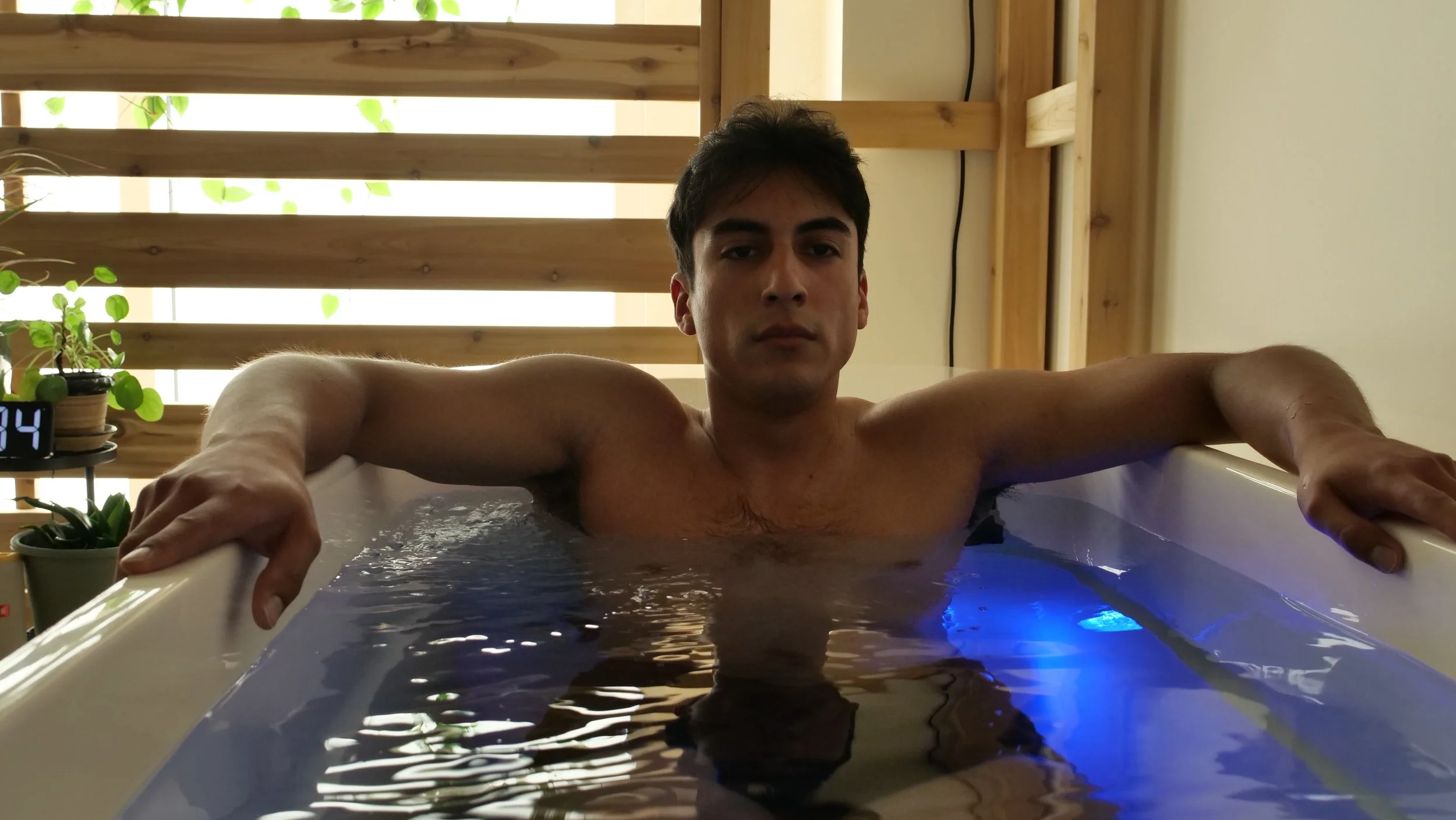 A young man with dark hair sitting in a cold plunge filled with water, with a cedar wood structure and plants in the background.