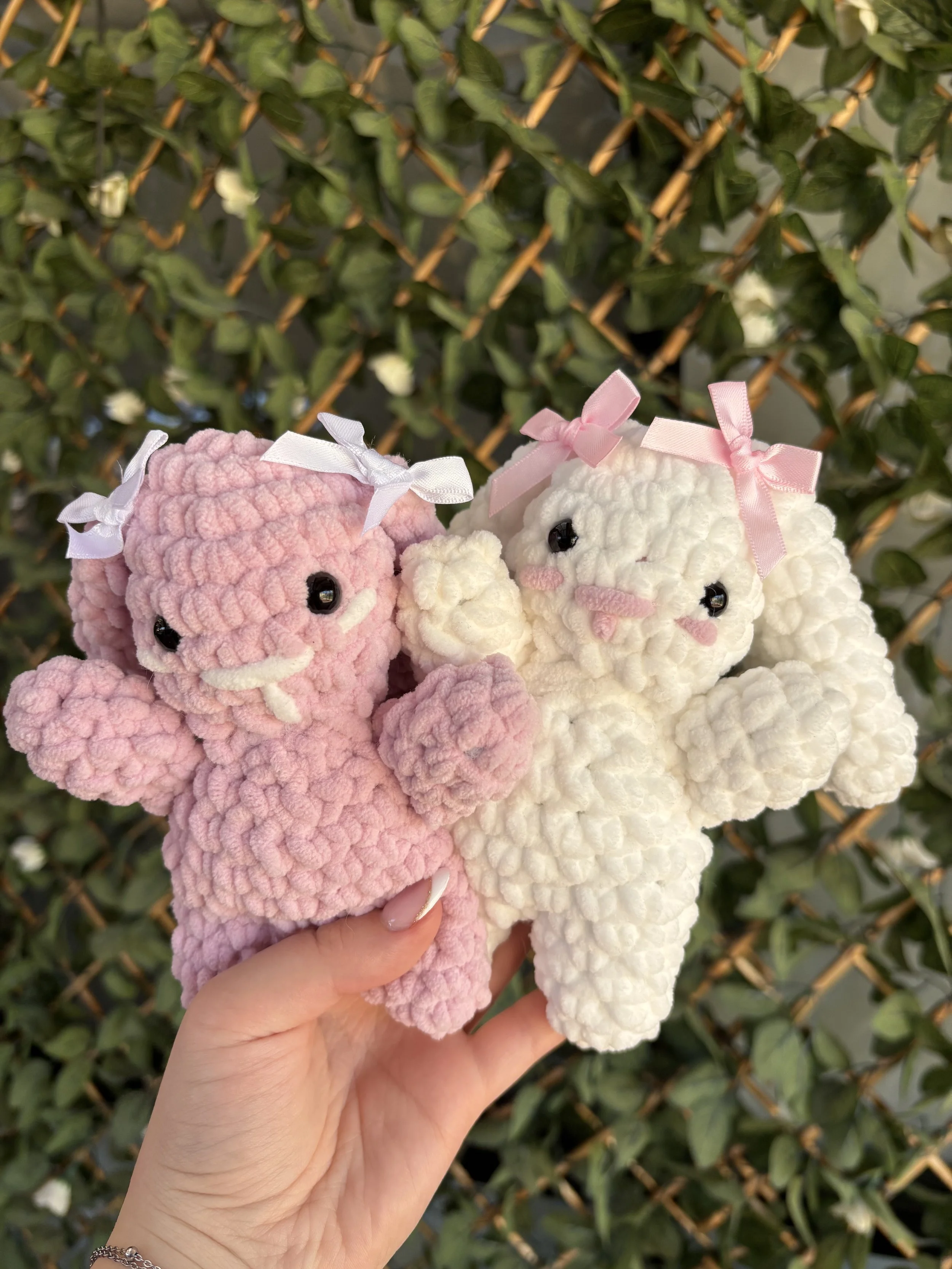 A hand holding three plush lamb toys, two white with pink ribbons and one pink with a white ribbon, in front of a green leafy background.
