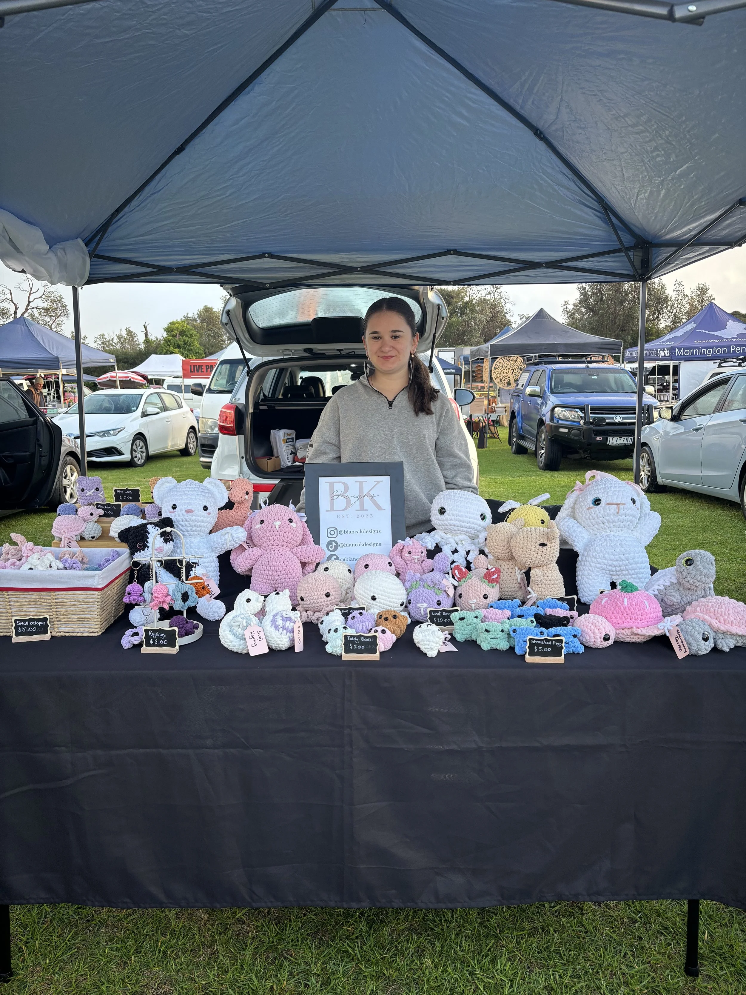A woman standing behind a table of handmade crocheted plush toys and keychains at an outdoor market stall, with tents and cars in the background.