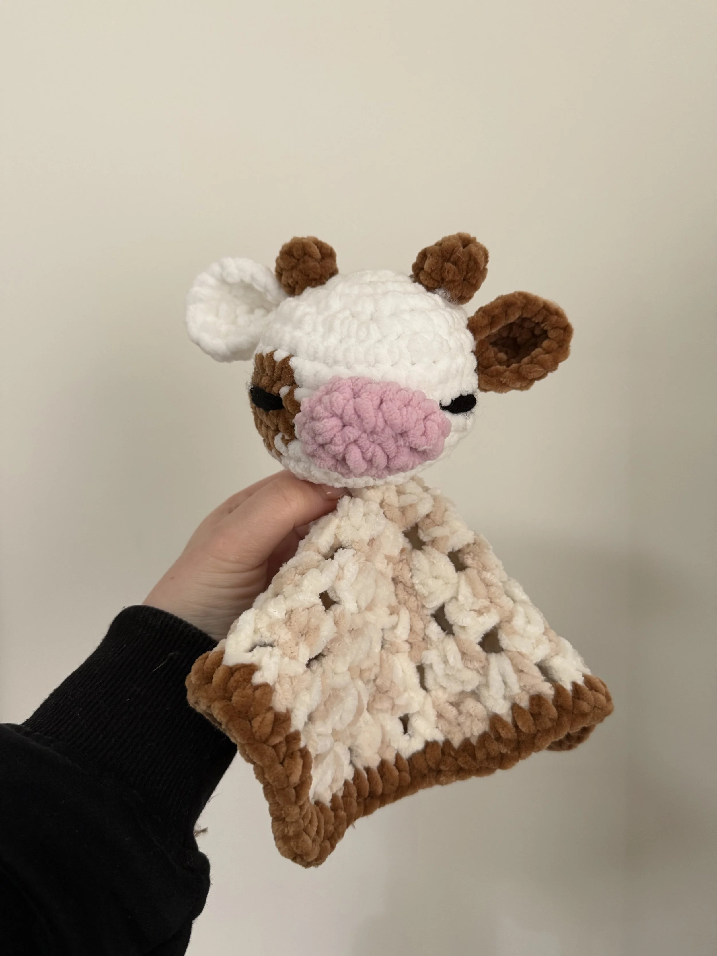 A handmade crocheted plush toy of a giraffe, with a beige body, brown spots, pink nose, and small brown horns.