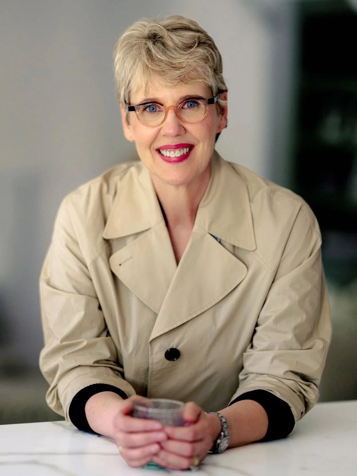 Barb Glass, an Intuitive strategy coach in WA, with short gray hair, wearing glasses and a beige trench coat, smiling and holding a cup of coffee, sitting at a table with a blurred background.