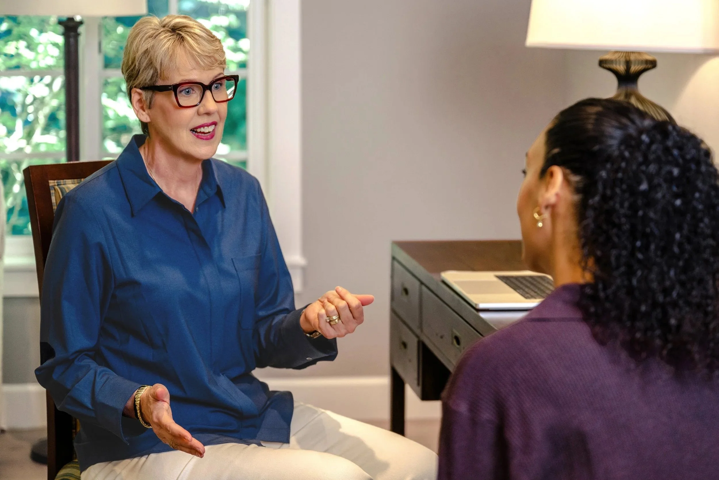 Two women are sitting and talking in a room with large windows. The woman on the left has short blonde hair, glasses, and is wearing a blue shirt. She is smiling and gesturing with her hands. The woman on the right has dark, curly hair, and is wearing a purple top. They are engaged in a conversation.