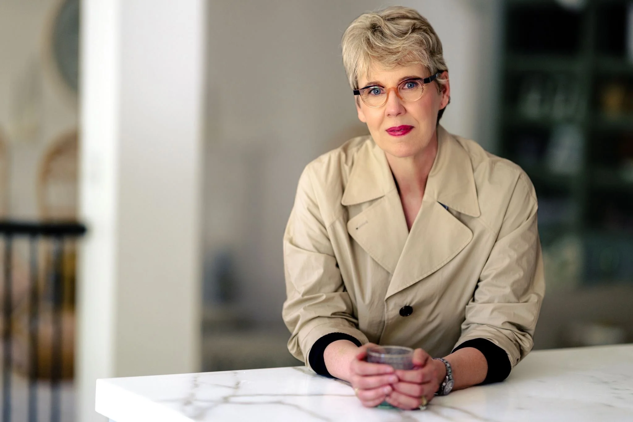 A woman with short blonde hair, glasses, and wearing a beige trench coat, sitting at a white table holding a glass of red wine, looking towards the camera, in a modern indoor setting.