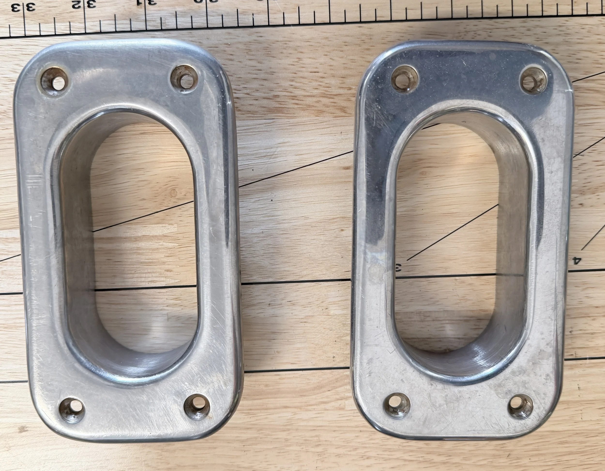 Two metal wall plates with four screw holes each and a large oval opening in the center, placed on a wooden surface.