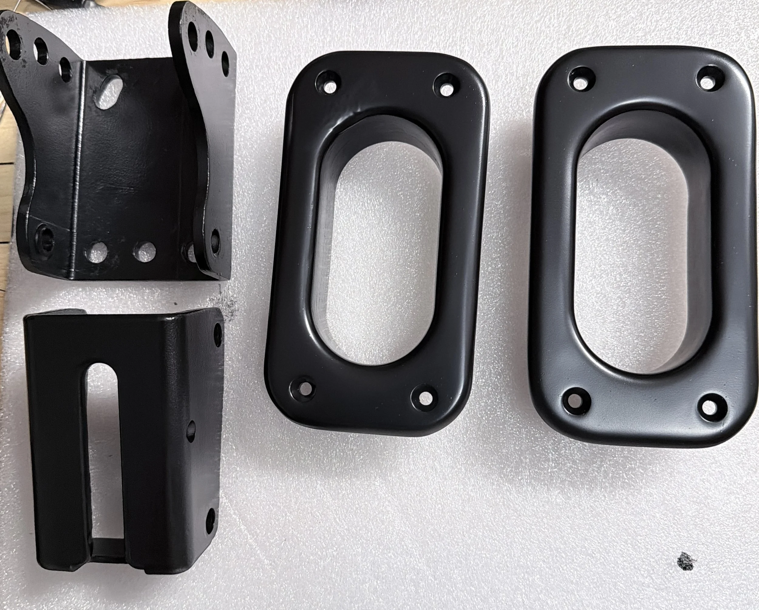 Three black metallic mounting brackets with multiple holes, placed on a light-colored surface; the brackets vary in shape and size.
