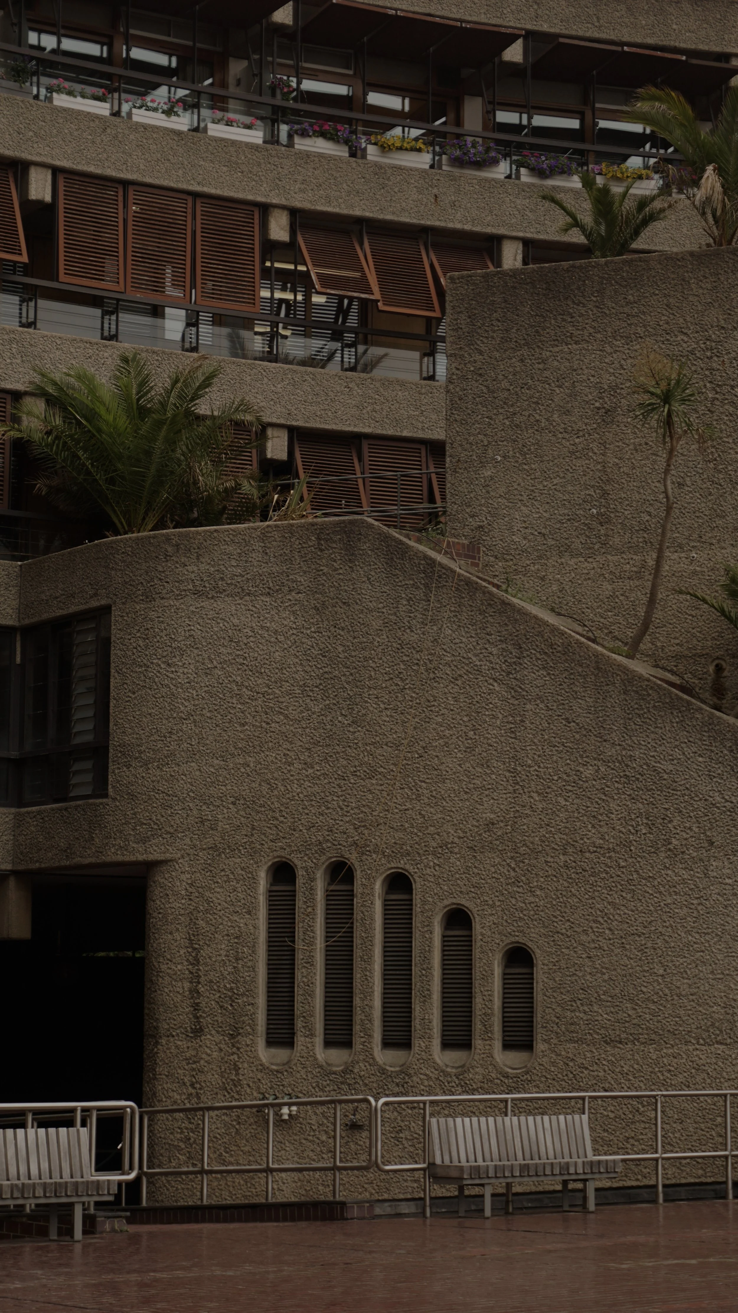 The image shows the exterior of a modern building with textured concrete walls, wooden window shutters, and balconies adorned with colorful flowers. There are palm trees and benches in the foreground.