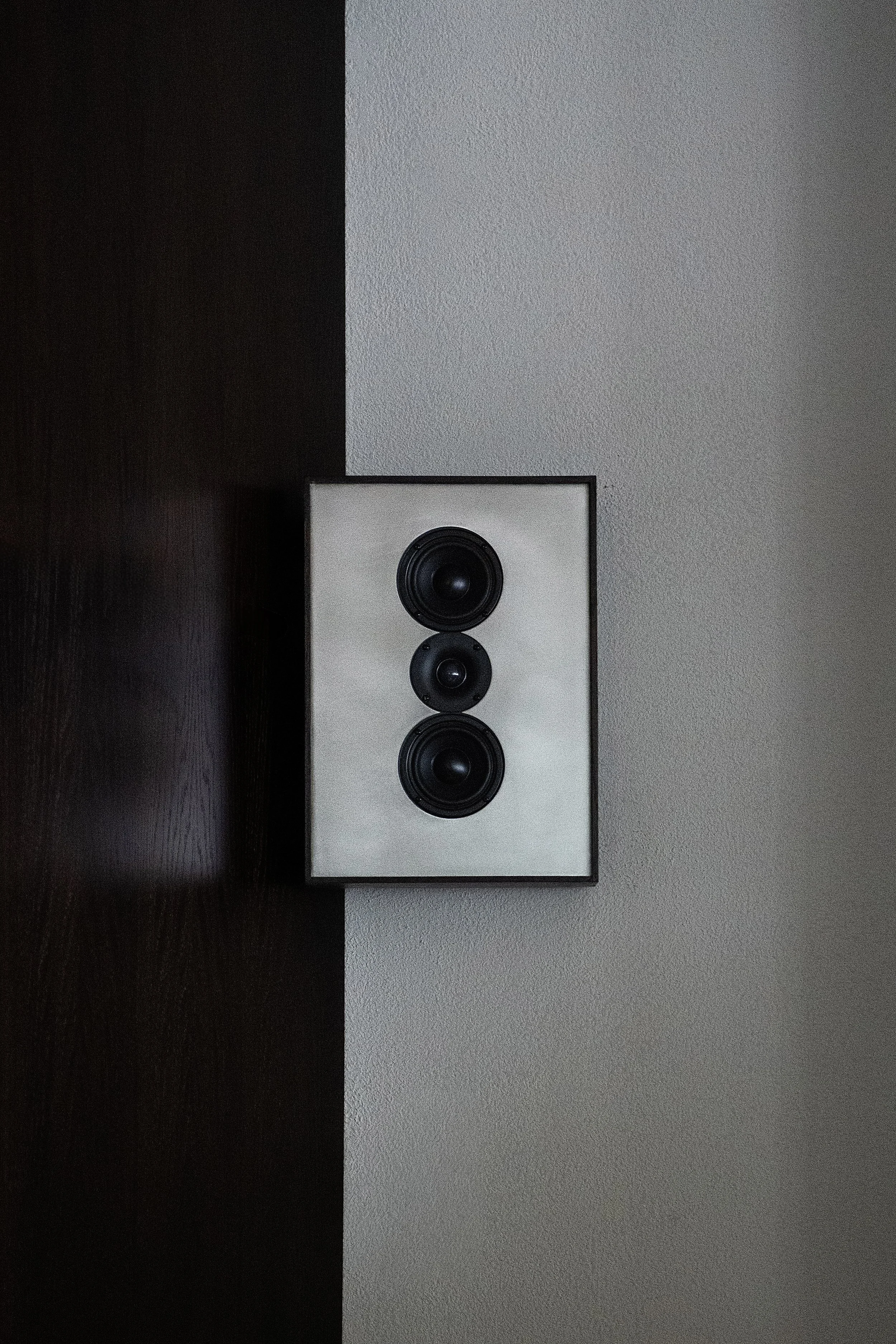 A white speaker with three black drivers mounted on a dark brown wooden surface, against a textured light gray wall.