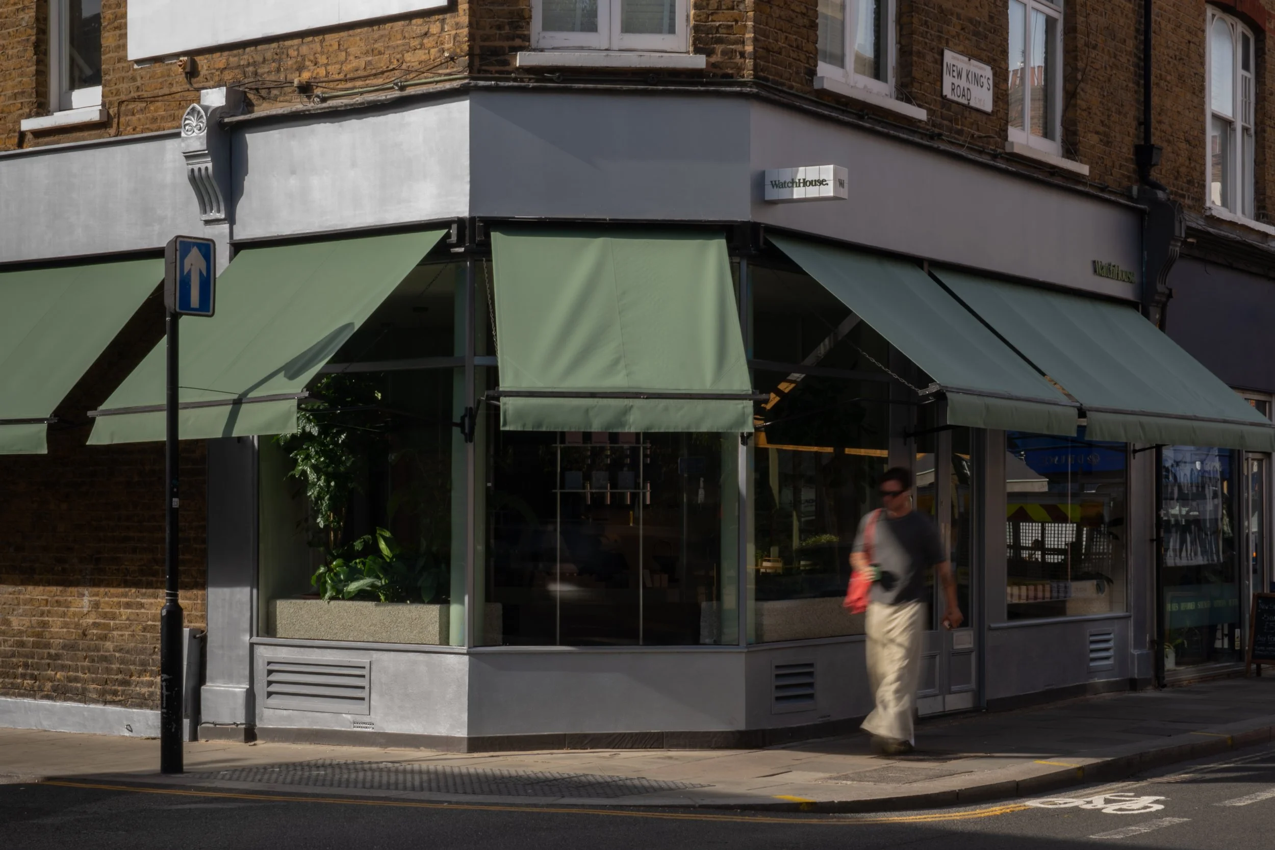 A street corner with a small storefront named WatchHouse, featuring green awnings and a glass window display. A street sign indicates the corner of New King's Road. A person is walking past the storefront.