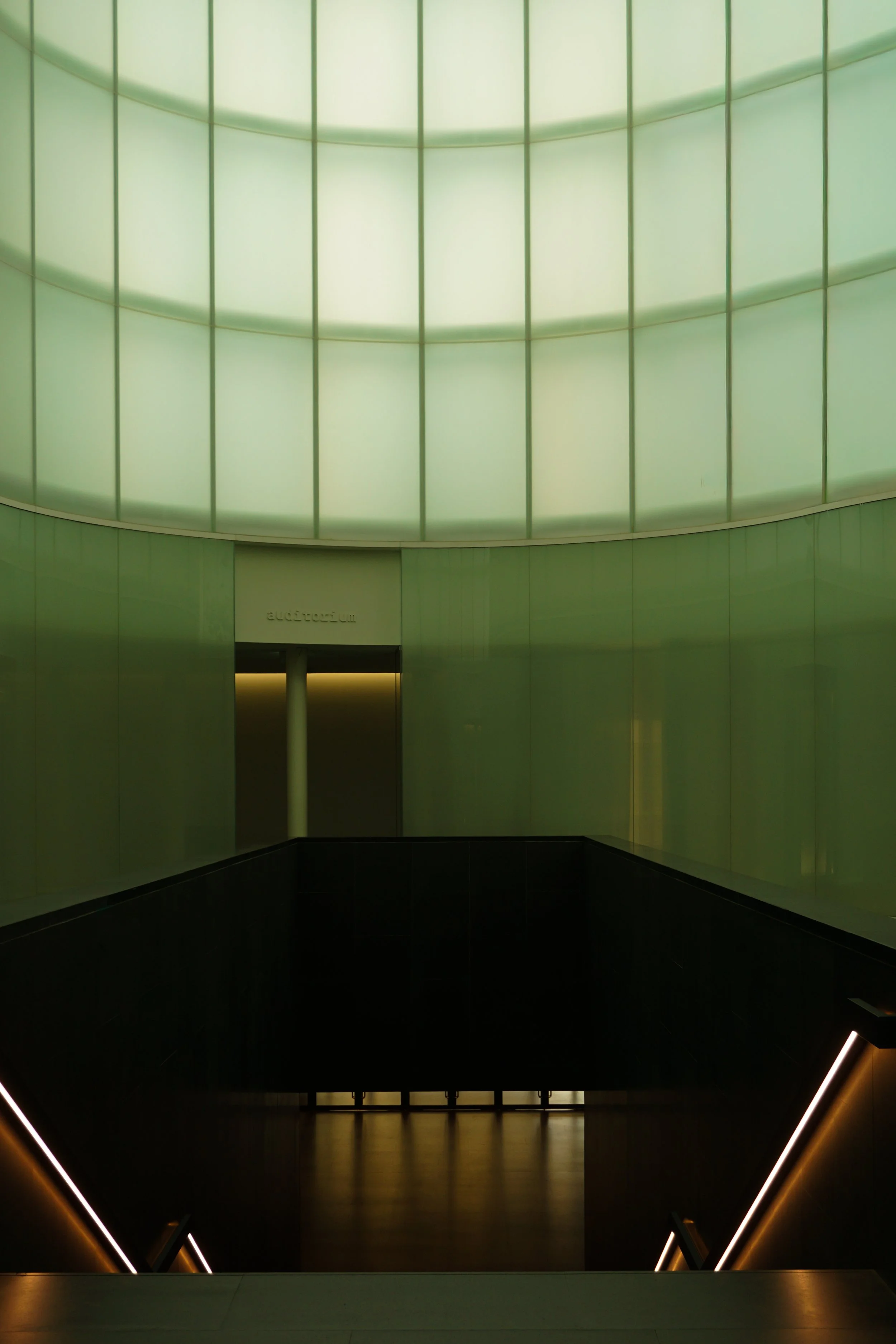 A modern indoor architectural space with soft green lighting and black flooring, featuring curved walls and minimal decorative elements.