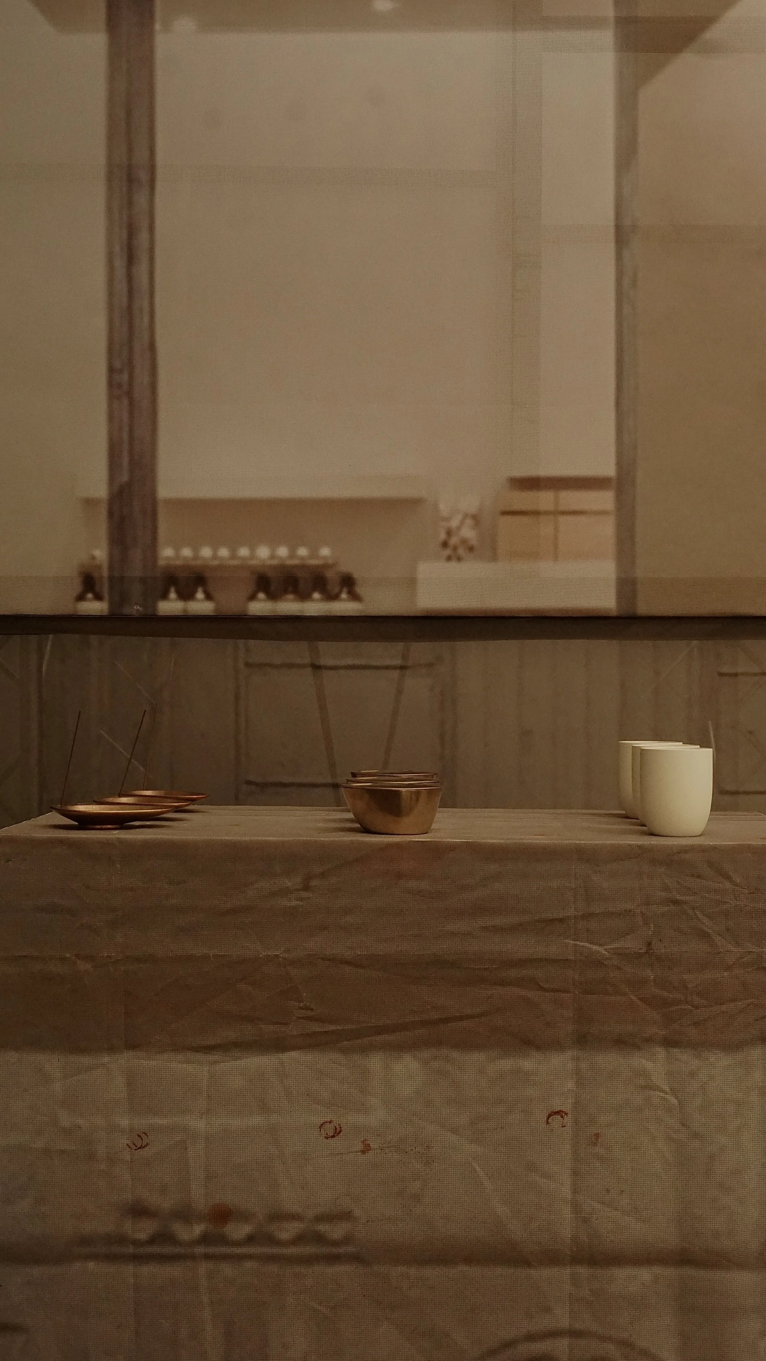 A minimalist table setting with two small alternating bowls, a larger central bowl, three small cups, and three small saucers, viewed through a window or glass partition.