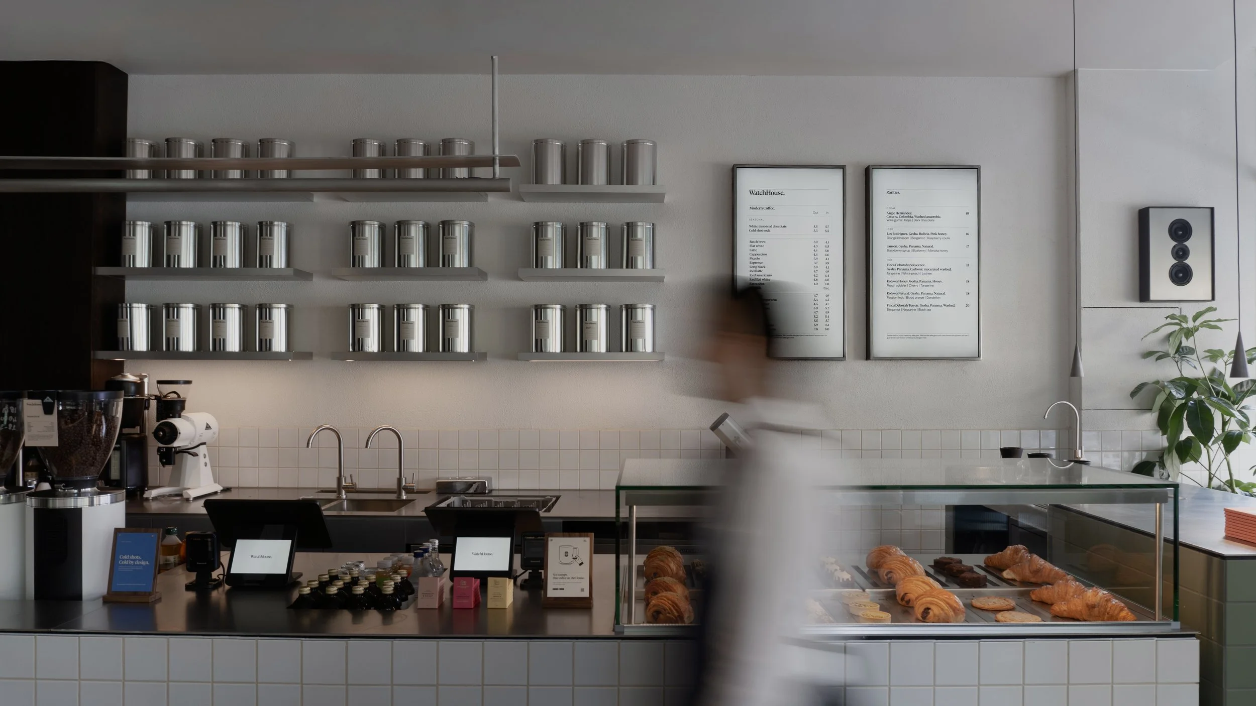 Coffee shop counter with shelves of coffee canisters, a coffee grinder, a pastry display with croissants and chocolate treats, and a menu on the wall.