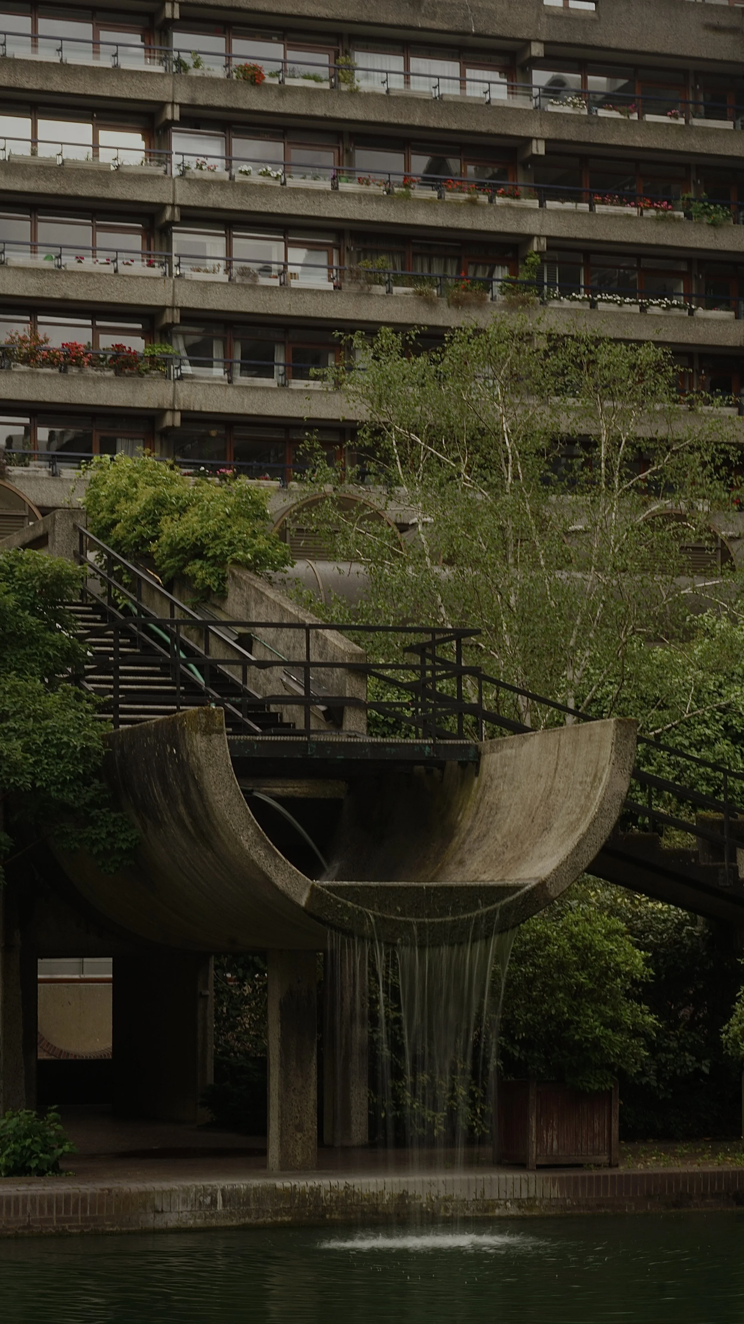 Apartment building with balconies decorated with plants and flowers, and a water fountain in front.