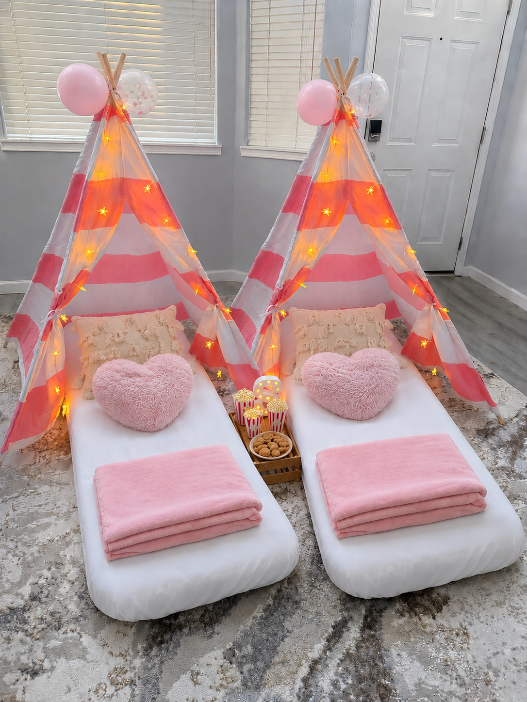 Two indoor beds with pink and cream bedding, decorated with plush heart-shaped pillows, topped with pink fleece blankets, and each placed inside a pink-and-white striped tent. The tents are adorned with yellow star-shaped string lights and topped with balloons and a small decorative teepee. A small wooden tray with popcorn and desserts is placed between the beds.