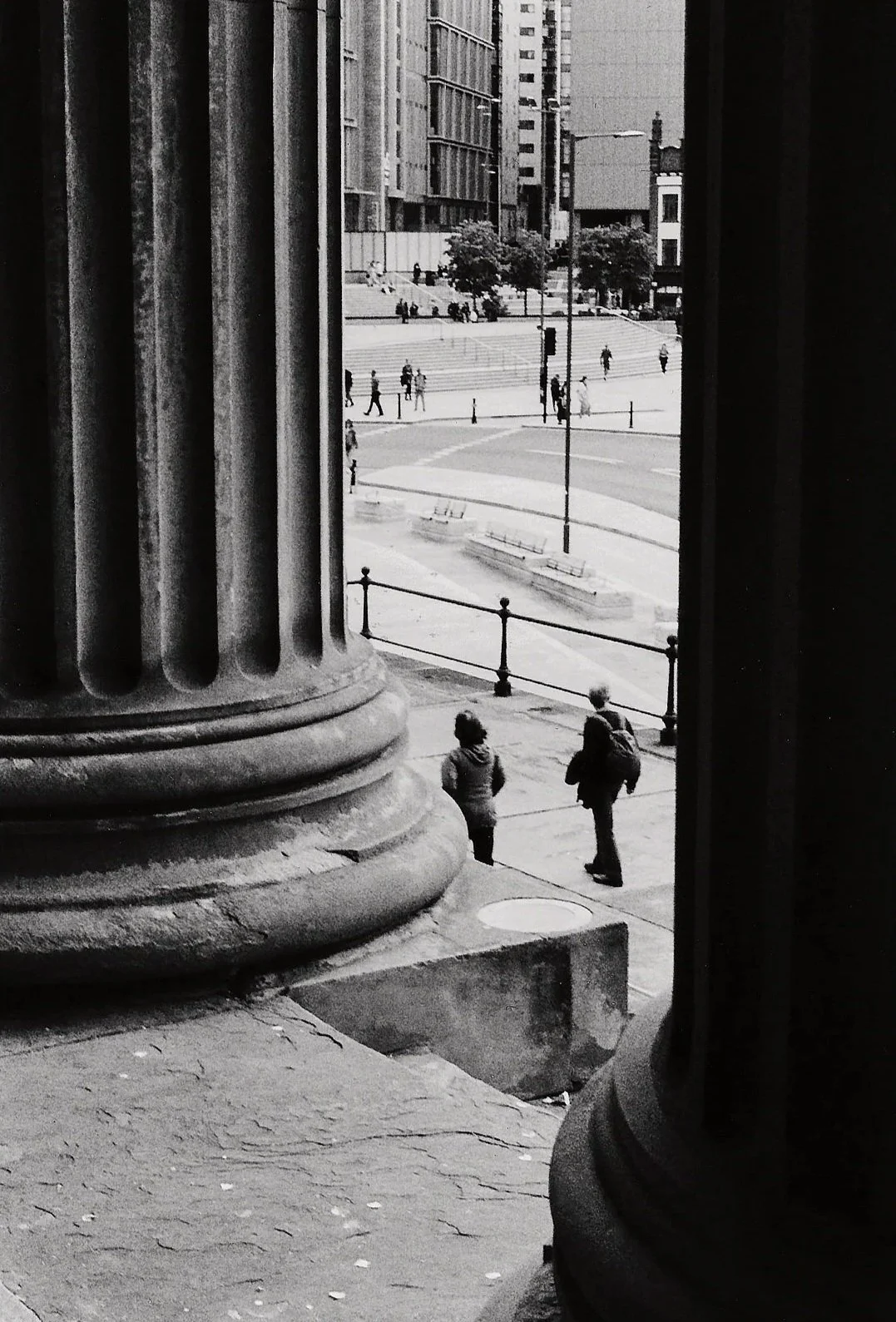 View of a city street through the columns of a historic building, showing pedestrians walking along the sidewalk and steps leading to an open plaza lined with trees.