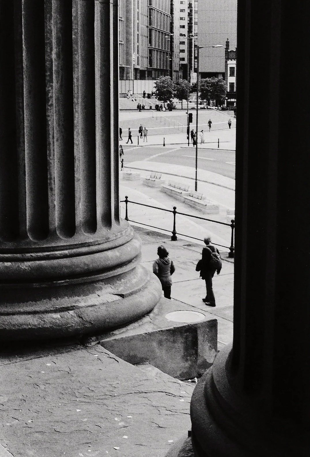 Black-and-white photo taken from inside a building showing a city street with pedestrians walking and stairs leading up to more people, with tall buildings and trees in the background.