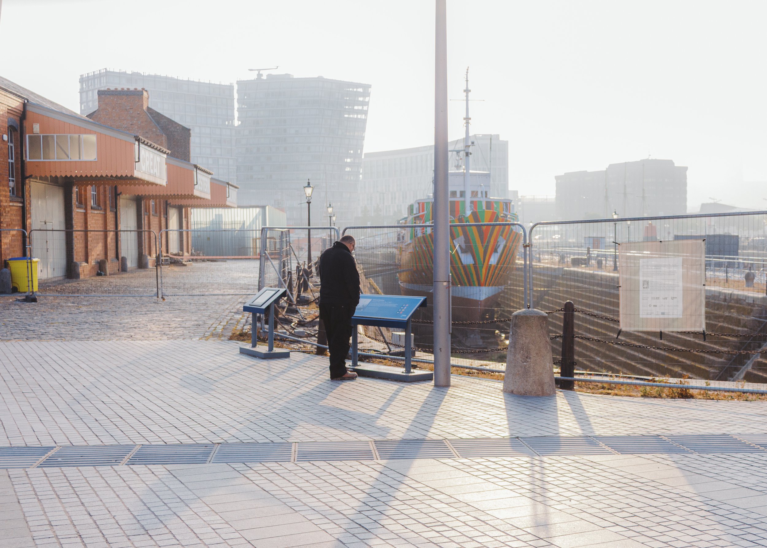 A man in a black coat standing outdoors near a fenced construction site with a colorful boat, old brick buildings, and modern glass skyscrapers in the background, under bright sunlight.