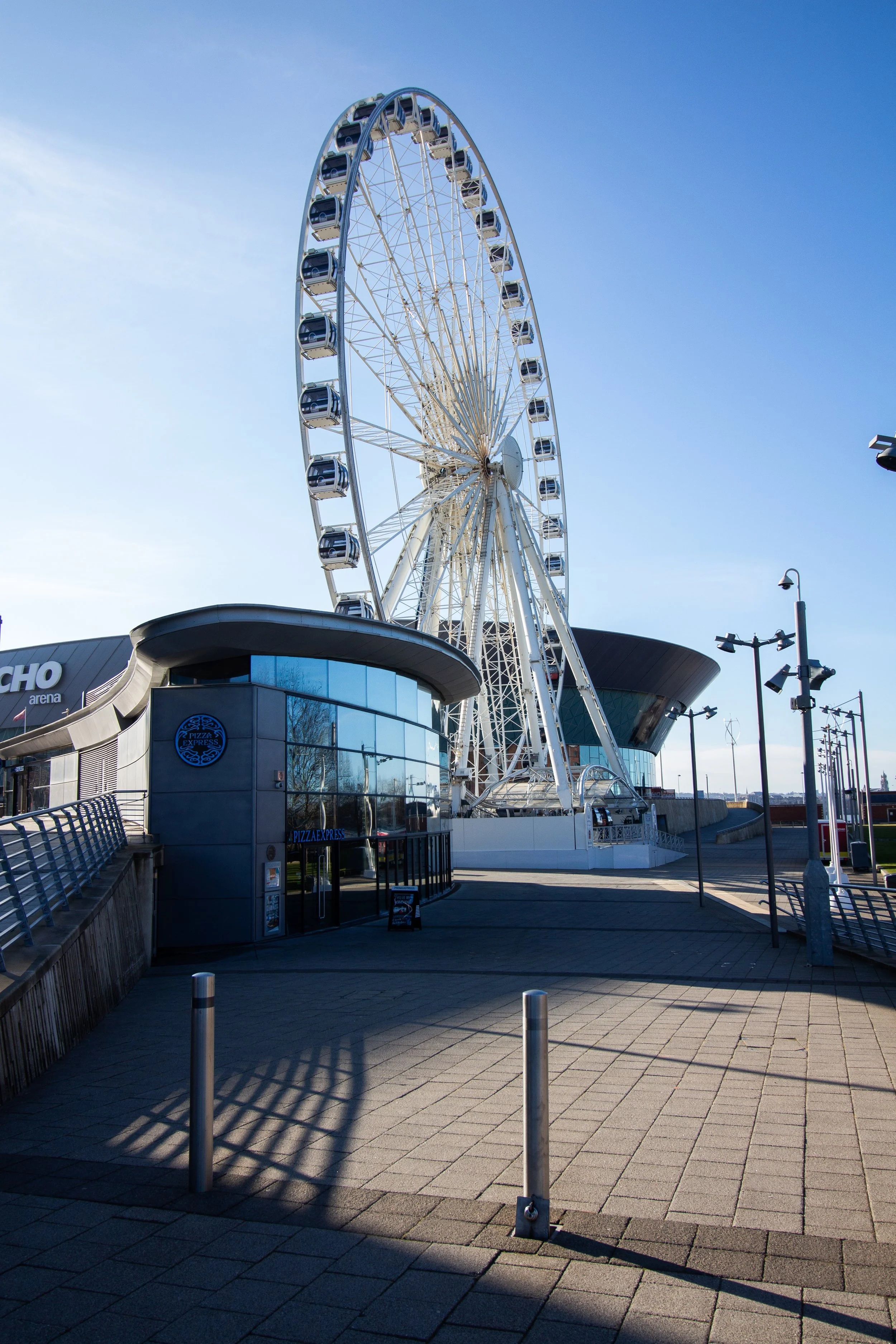 A large Ferris wheel outdoors near a modern building with a Pizza Express sign, under a clear blue sky. Street lamps and metal barriers are visible in the foreground.