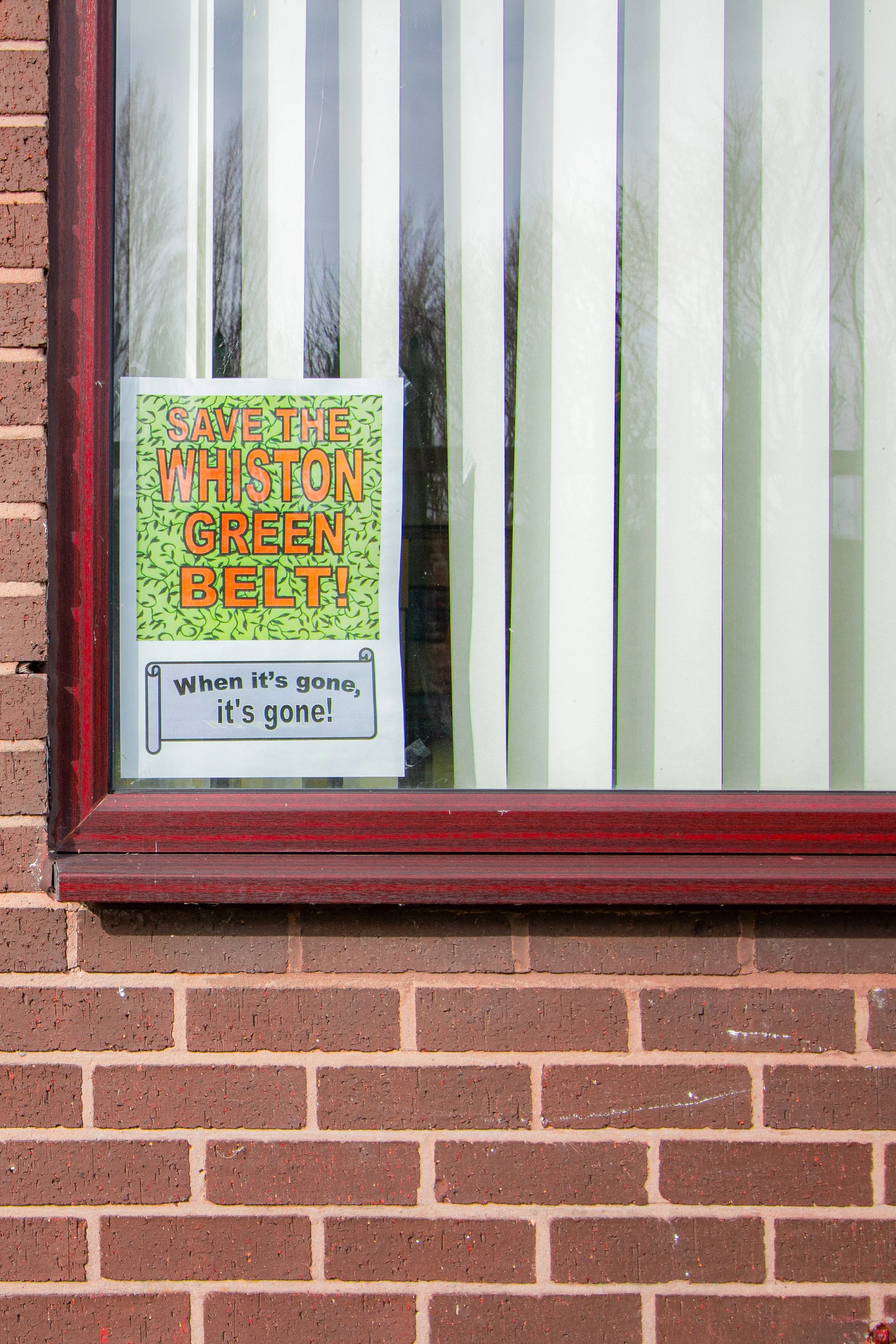 A window with white vertical blinds and a colorful sign that reads 'SAVE THE WHISTON GREEN BELT!' attached to the glass. The sign has a green leafy background and orange text. The window is set in a brick wall, and there is a reflection of trees in t