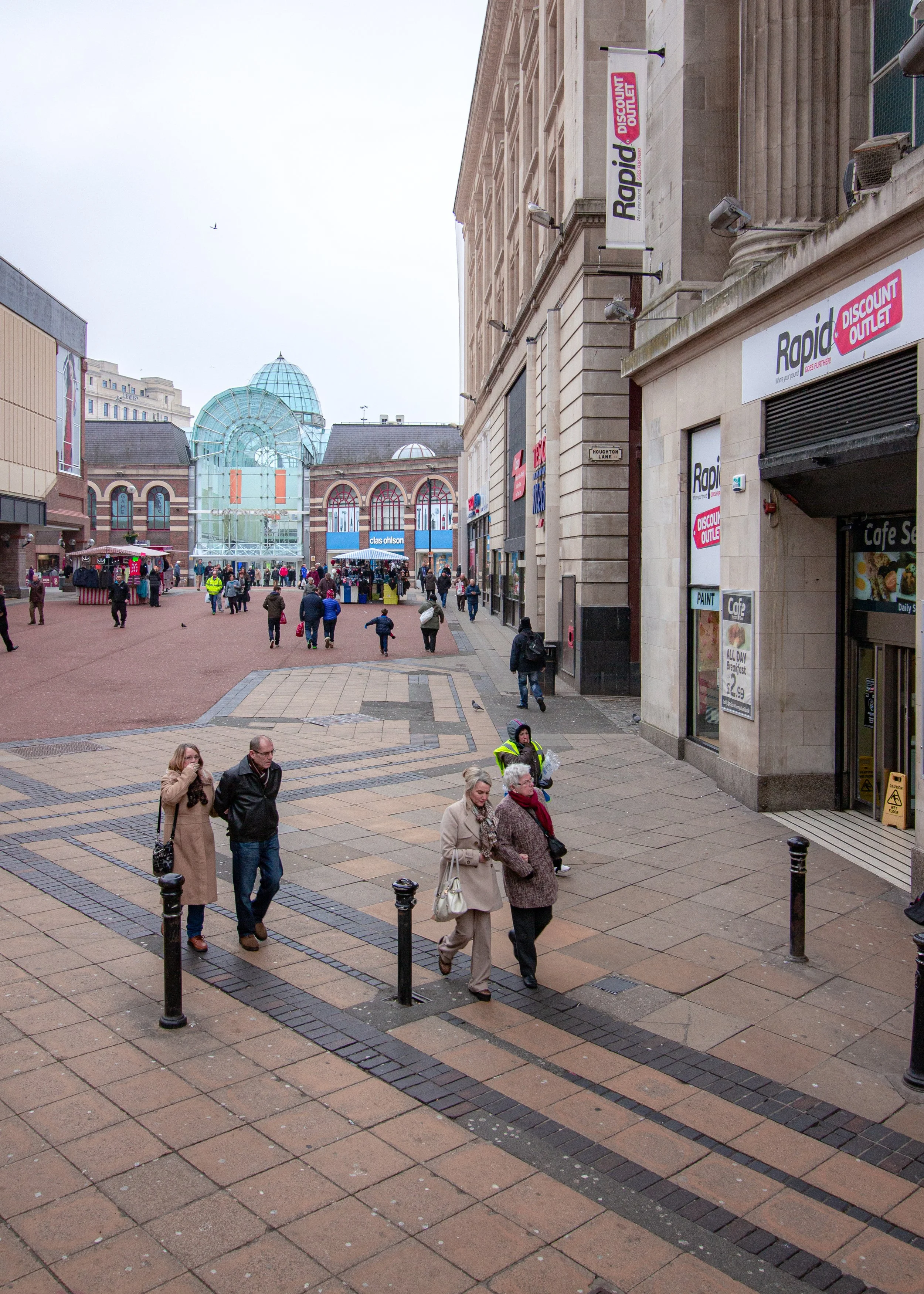 City street scene with pedestrians walking and shopping on a cloudy day. Buildings and storefronts line the street, with a large glass structure in the background.