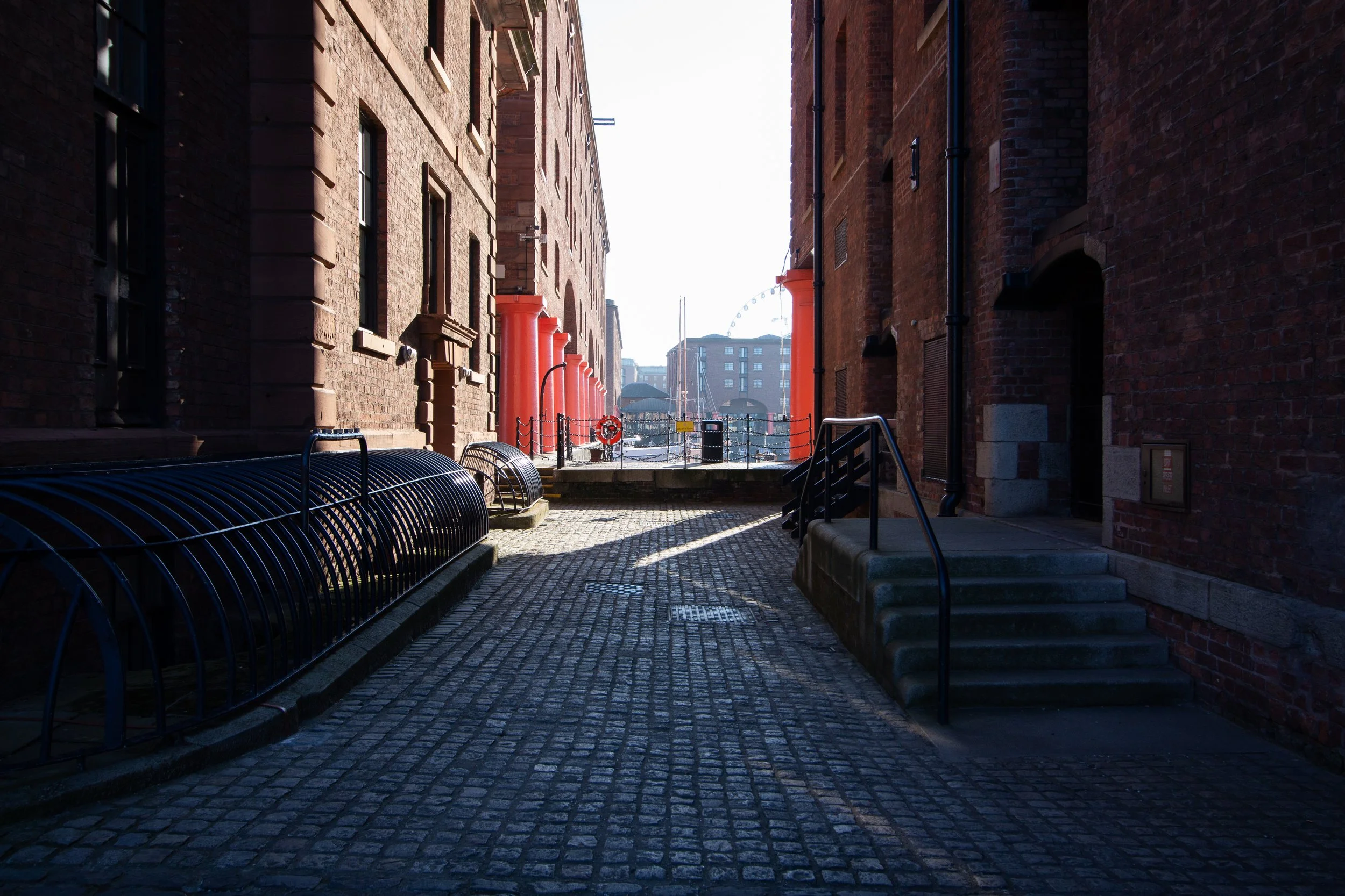Narrow alleyway between brick buildings with red columns, cobblestone pavement, and a view of a ferris wheel in the distance.