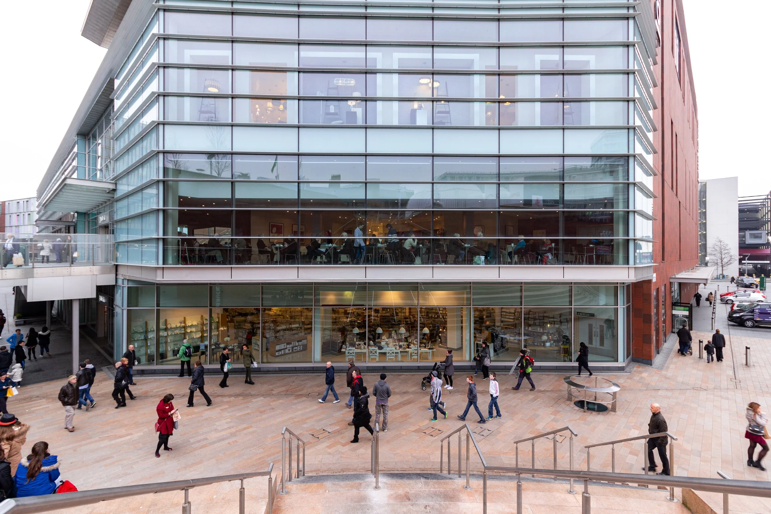 Modern multi-story building with large glass windows and a bookstore on the ground floor. People walking outside in a plaza with stairs and handrails.