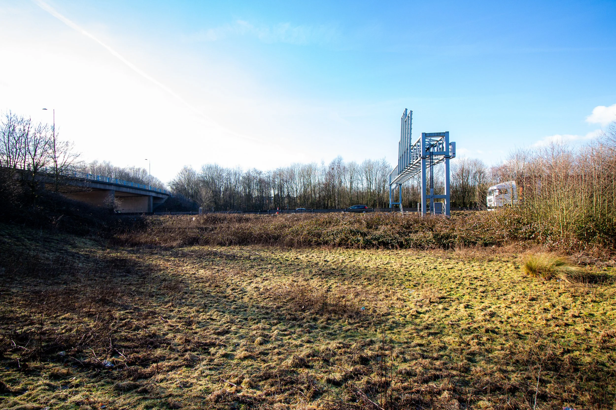 A landscape with a grassy foreground, a highway overpass on the left, and a billboard frame structure on the right against a bright blue sky with a few clouds.