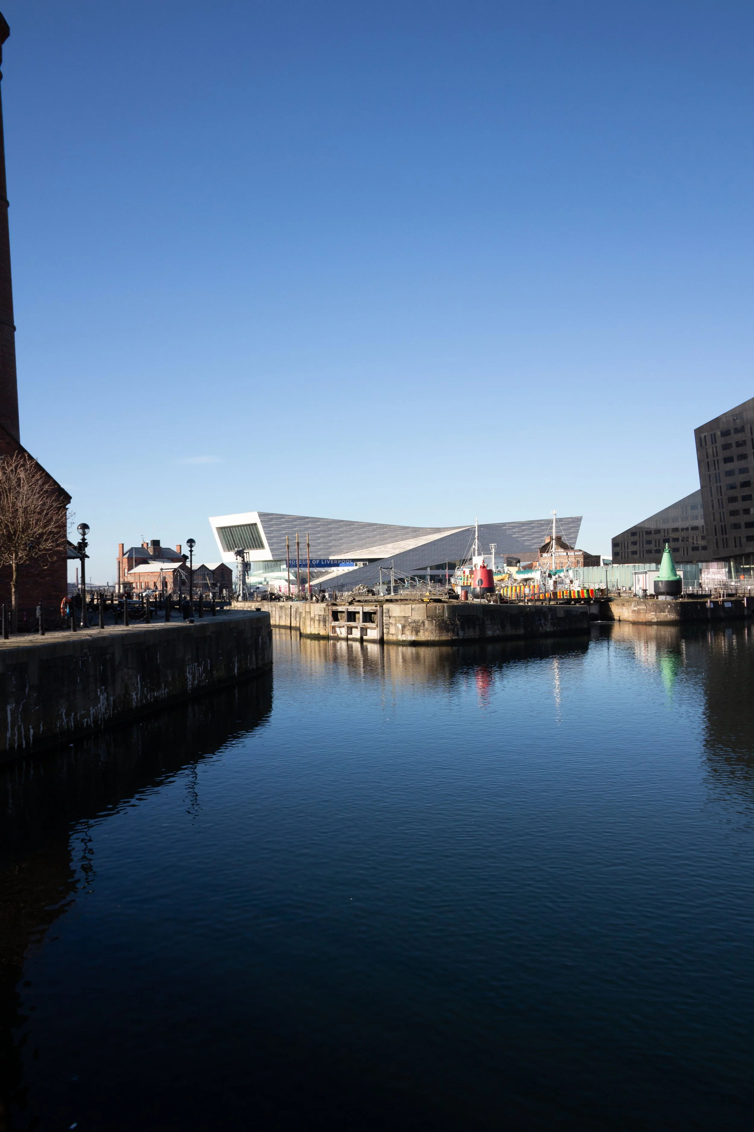 View of a river with dark water reflecting nearby buildings and boats, with a modern museum building in the background and clear blue sky.