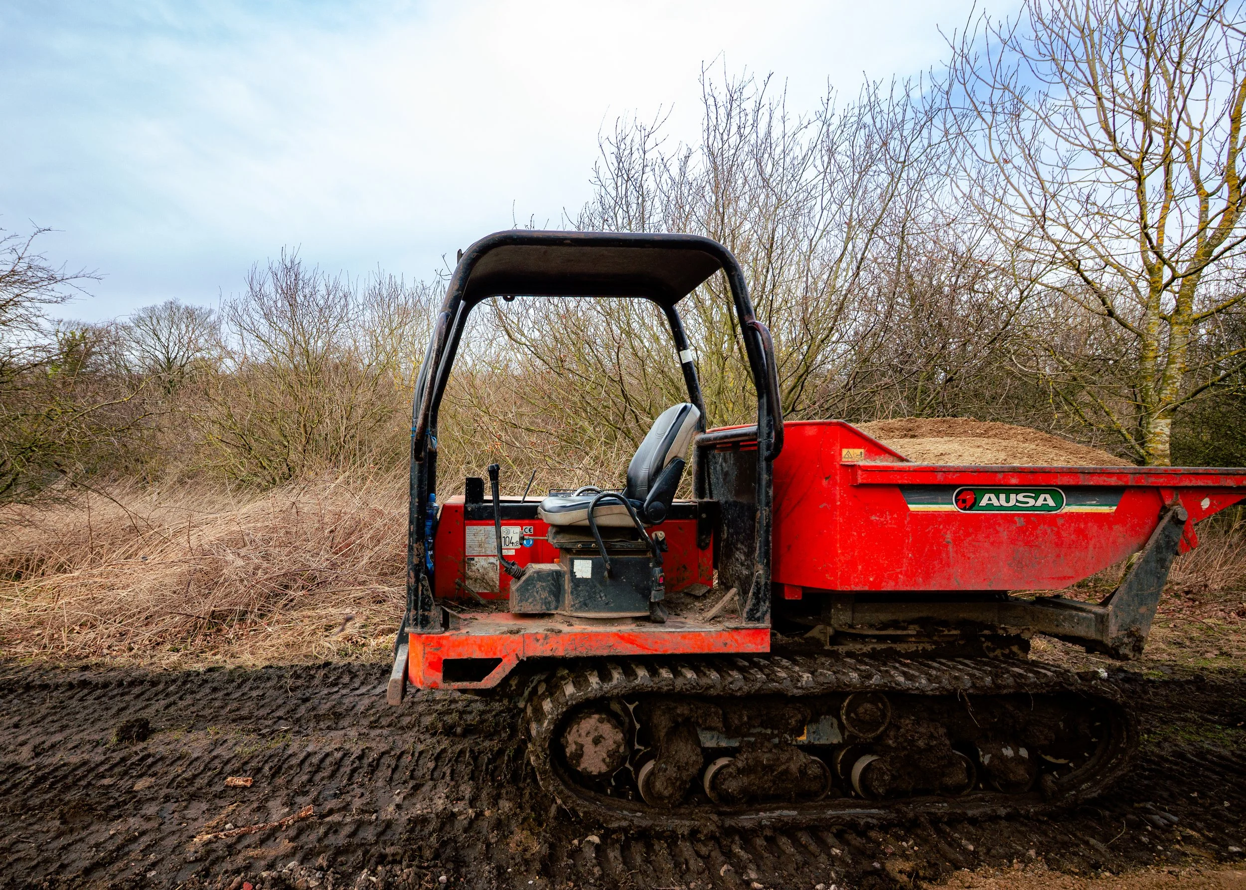 A red tracked utility vehicle with an open cabin, carrying dirt or mulch, on a muddy, rutted path in a wooded area with leafless trees and an overcast sky.