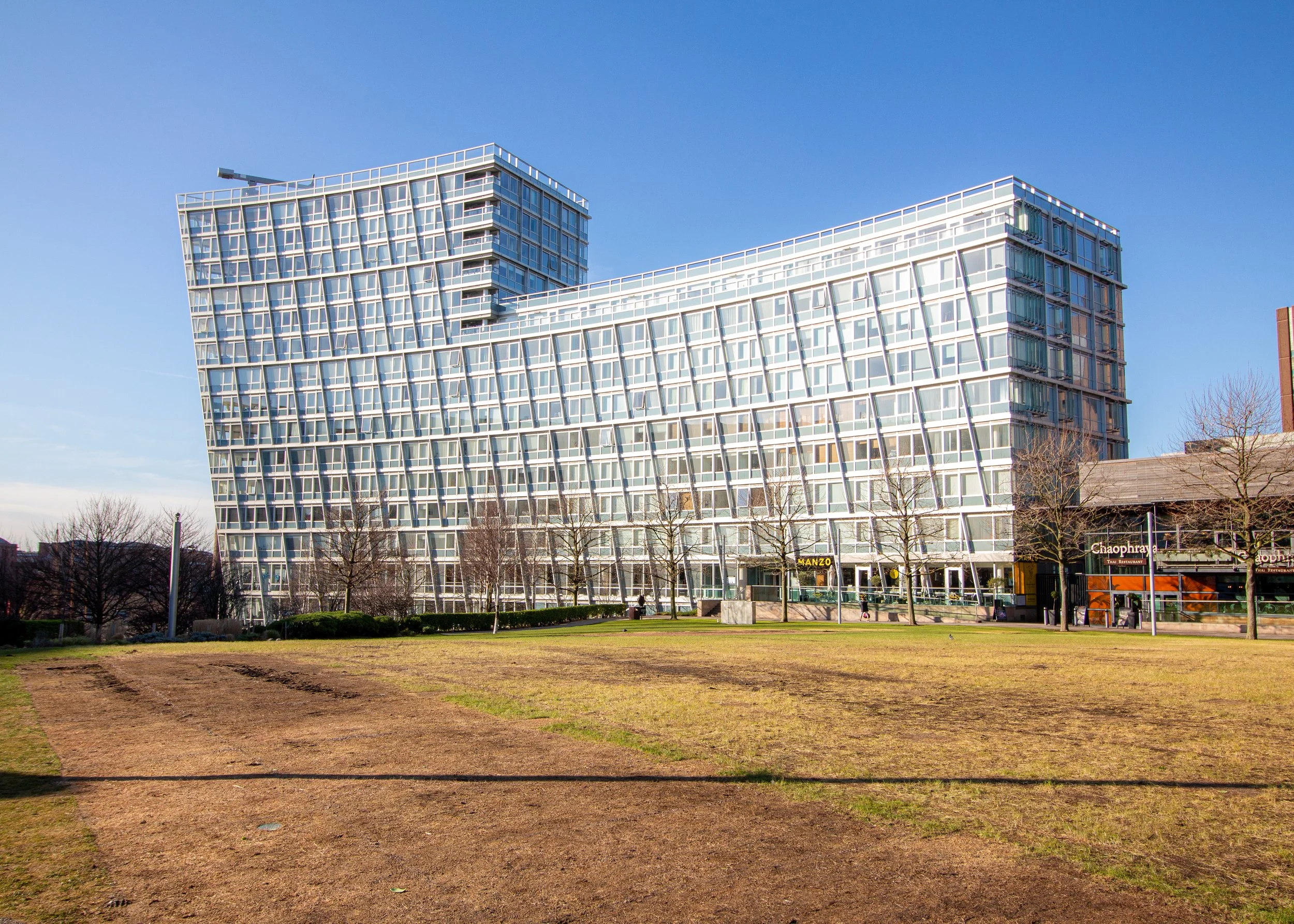 Modern glass building with unique angular architecture, seen from a park with a dirt path, leafless trees, and a clear blue sky.