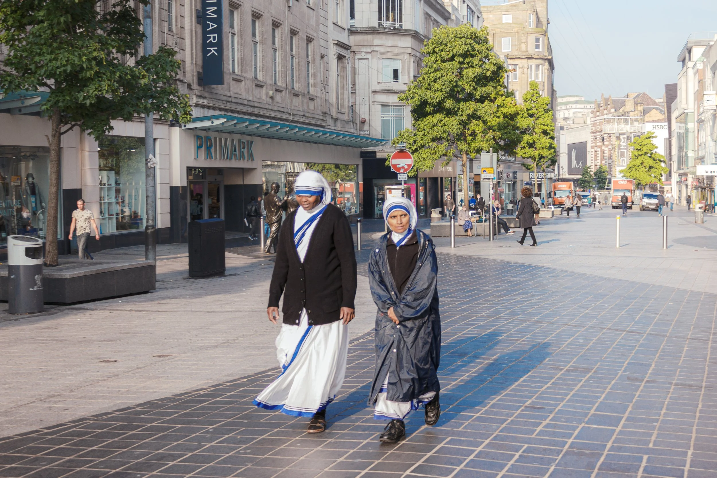 Two women dressed in traditional clothing walking on a city street with modern buildings, trees, and pedestrians in the background.