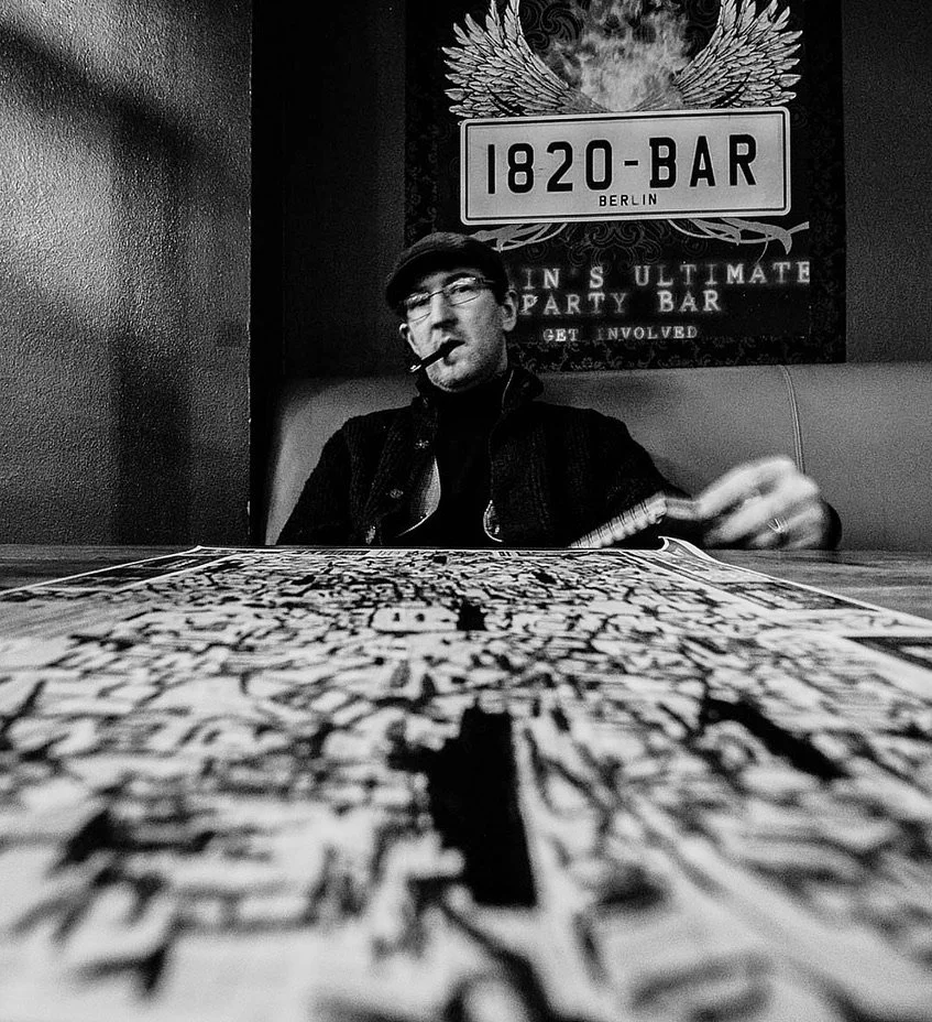 A man with glasses, wearing a hat and jacket, sitting at a table with a newspaper in front. He has a pipe in his mouth and is reaching across the table. Behind him is a sign that reads '1820-BAR Berlin' and a wall with a decorative design, including 