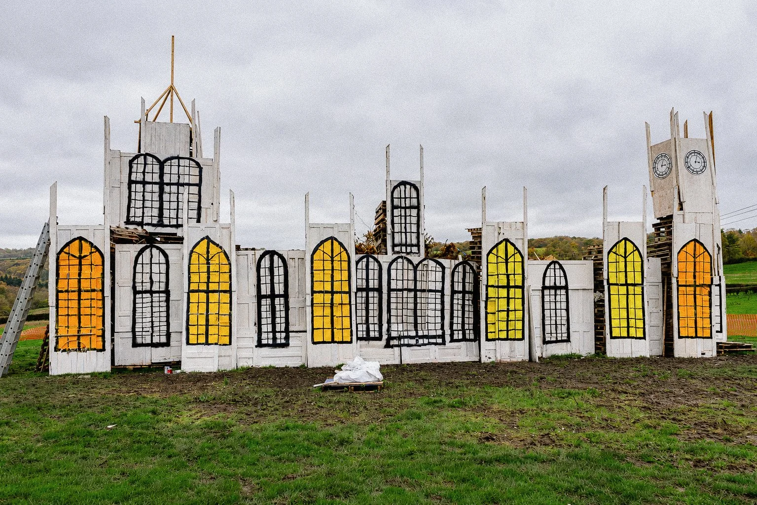 Partially constructed building facade with white walls and black window frames, decorated with yellow, orange, and gray panels, composed of wooden panels and structures, set outdoors in a grassy field under a cloudy sky.