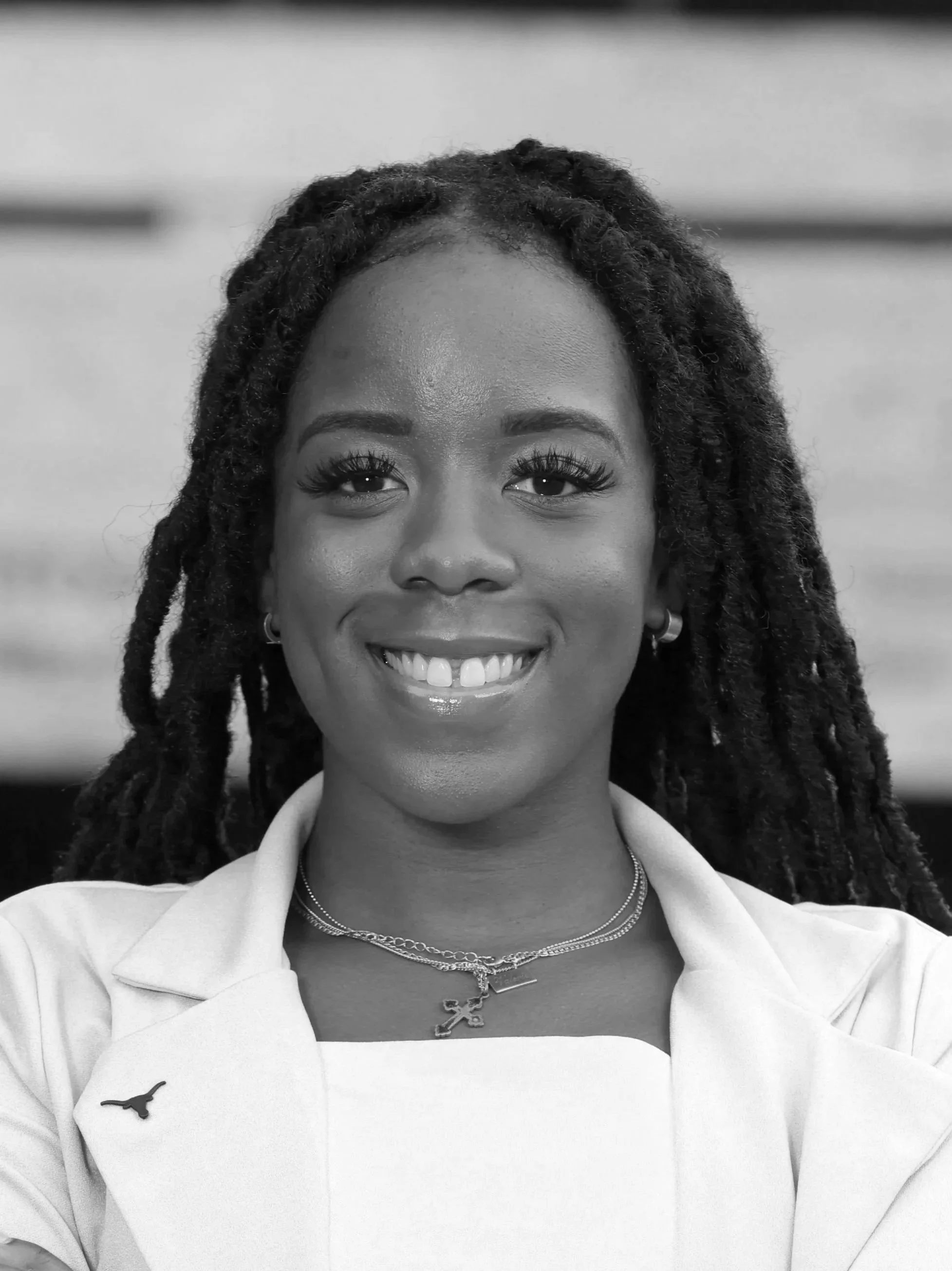 Close-up black-and-white photo of a smiling woman with long dreadlocks, wearing a blazer, jewelry, and a necklace with a cross pendant.