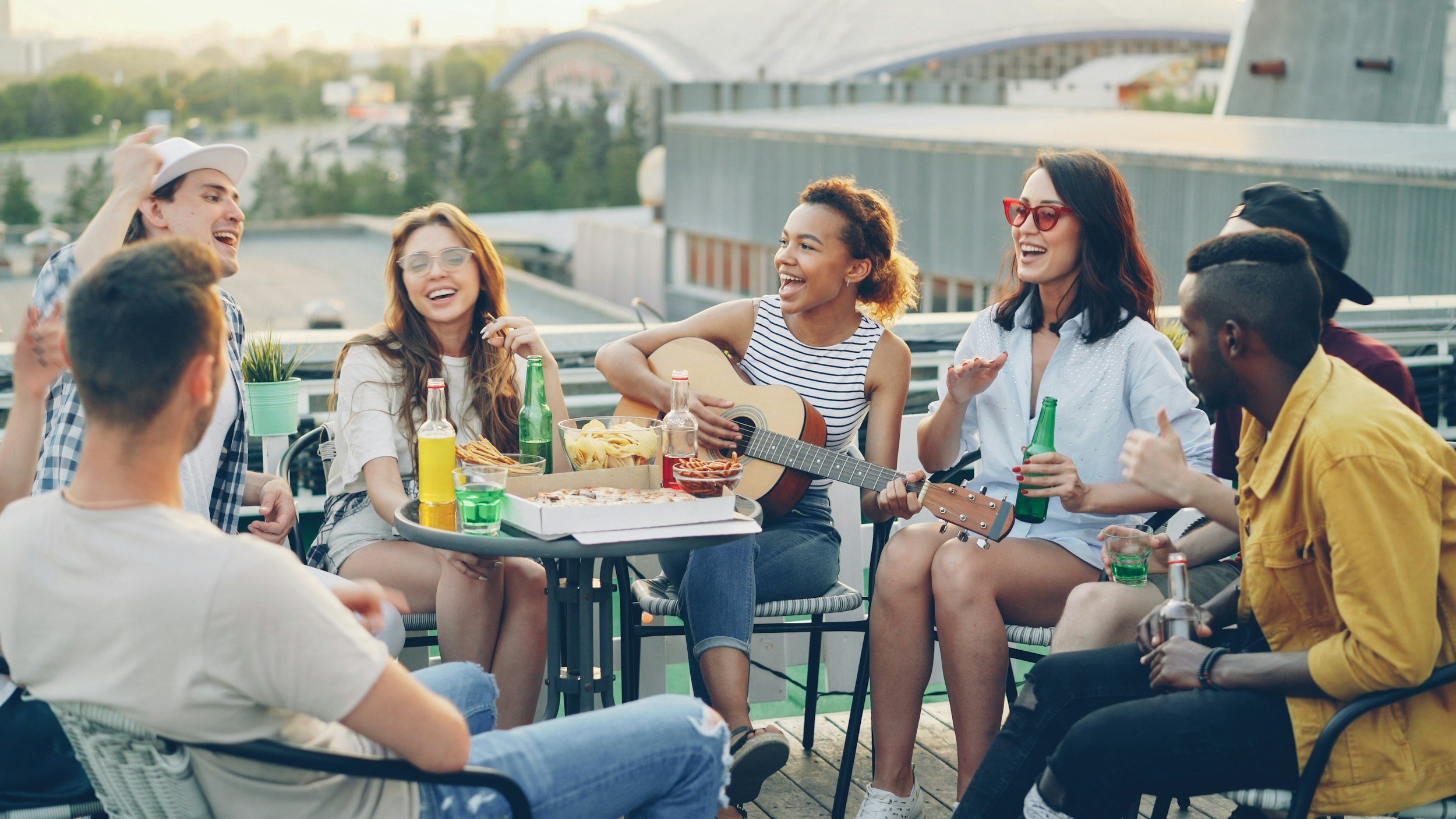 Group of young adults enjoying a rooftop gathering with food and drinks, some playing guitar and laughing in the evening sun.