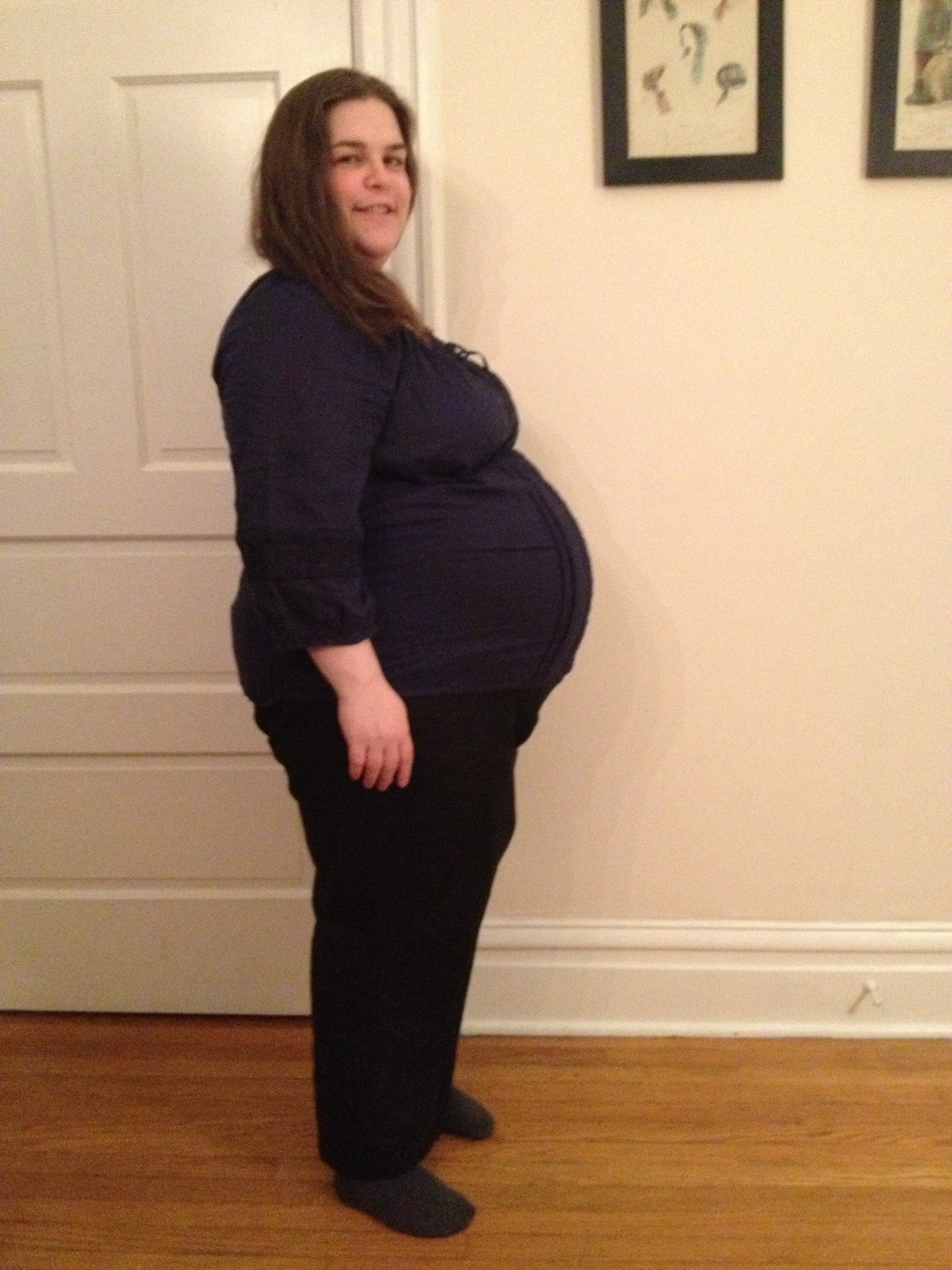 A pregnant woman with shoulder-length brown hair and wearing a dark long-sleeve top and black pants.