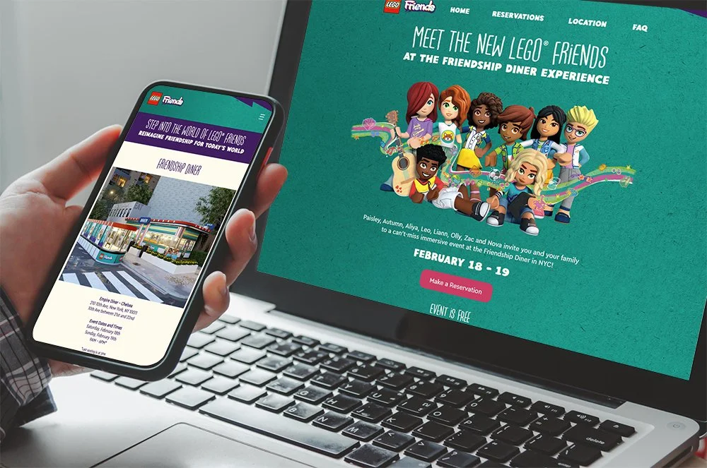 A person holds a smartphone displaying a webpage about LEGO Friends at a Friendship Diner event in NYC, with a laptop screen in the background showing a promotional webpage for the event and cartoon characters.