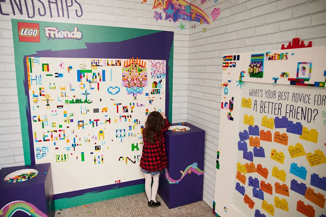 A young girl with long wavy hair, wearing a red and black checkered jacket and white tights, is placing LEGO bricks on a LEGO mosaic wall inside a colorful room. The wall features various LEGO pieces and seems to be a part of an interactive activity.