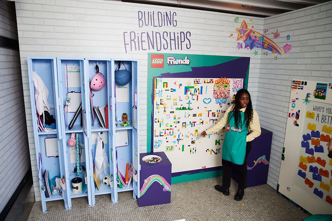 A girl in a teal apron stands in front of a whiteboard with pixel art, explaining or presenting. There are lockers filled with various items, and colorful posters and artwork on the brick wall that promote friendship and building relationships.