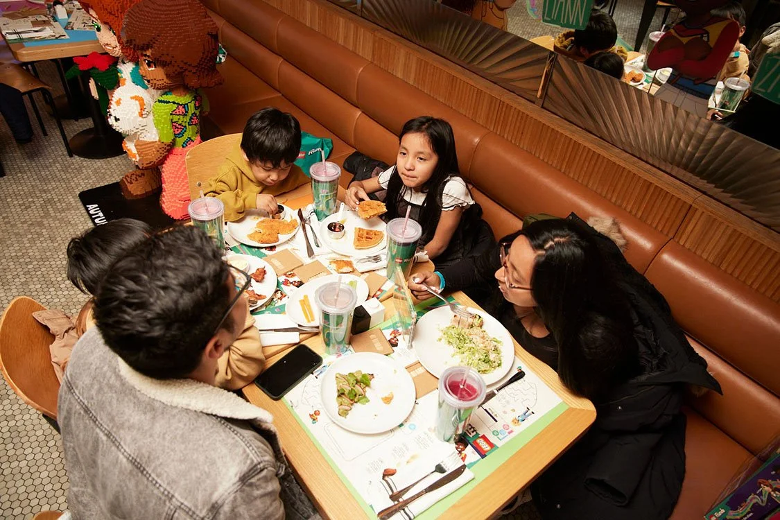A group of five people, three children and two adults, sit around a table at a restaurant, eating and drinking. They have plates of food including salad and chicken, and large drinks with straws. In the background, a colorful character figure made of