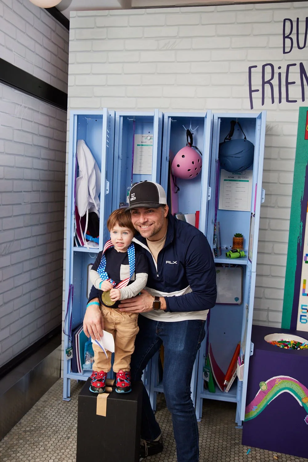 A man and a young boy smiling together in front of blue lockers. The boy is standing on a black box, holding a medal, and wearing a black jacket with patches, beige pants, red sneakers, and a medal around his neck. The man is crouching next to him, w