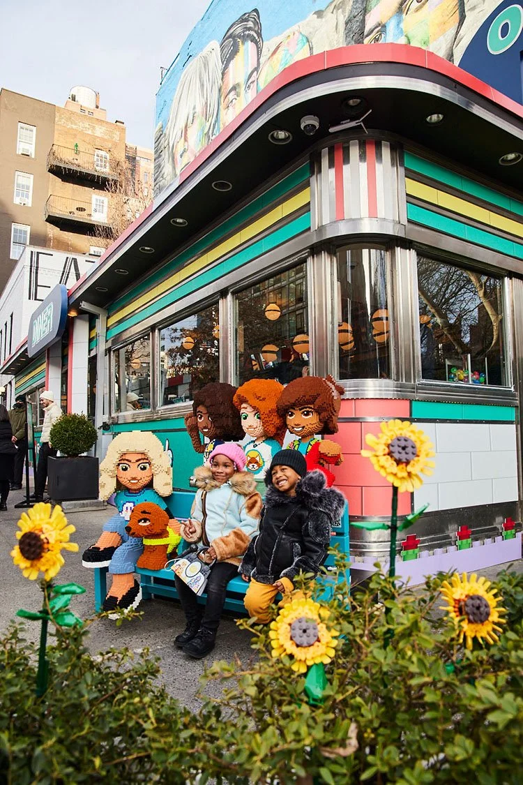 Three children sitting on a bench in front of the LEGO Friendship Diner with Lego characters and art, with sunflowers in the foreground.