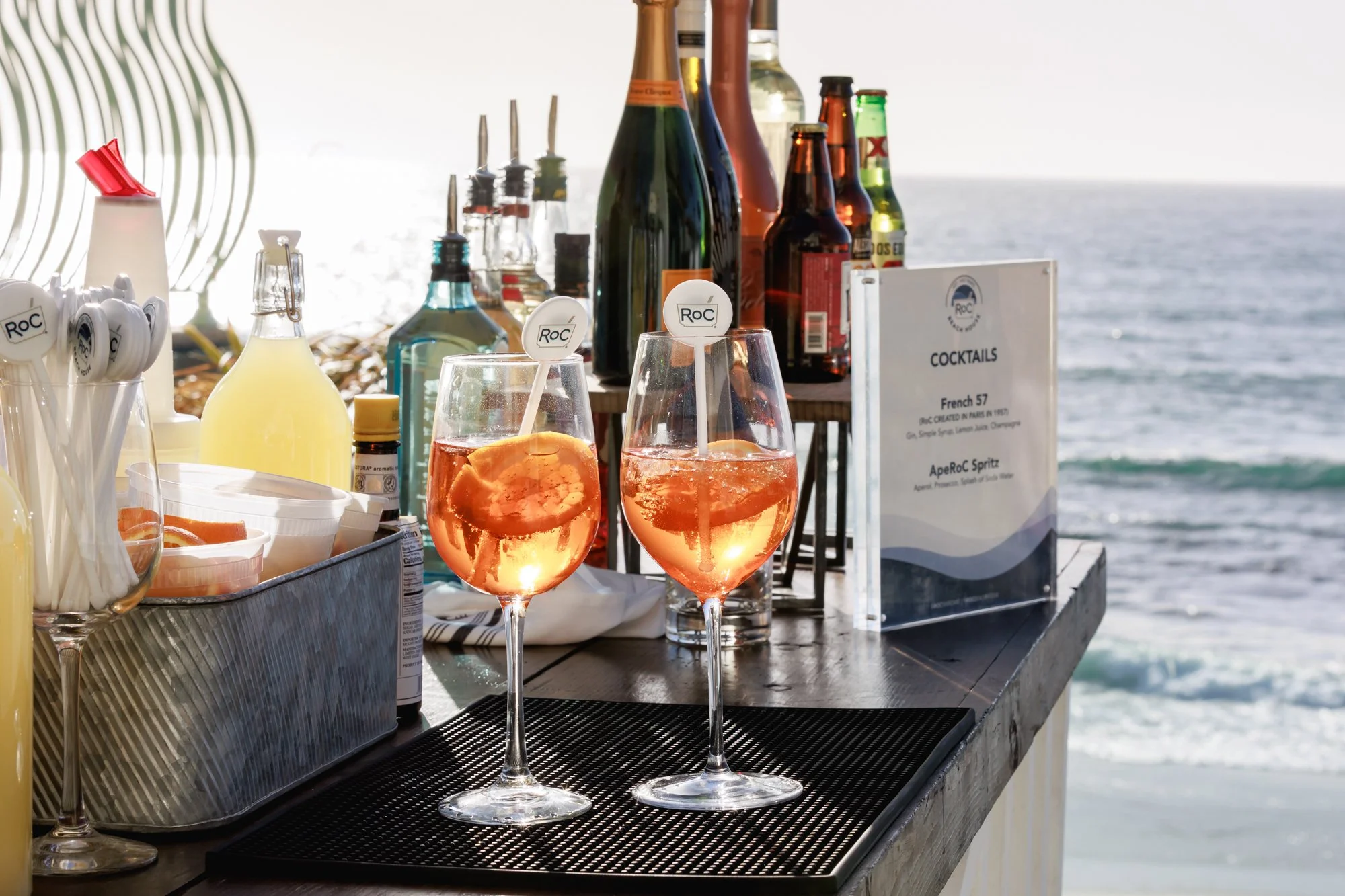 Two glasses of Aperol Spritz on a bar counter with a view of the ocean in the background, along with various bottles of alcohol and cocktail ingredients.