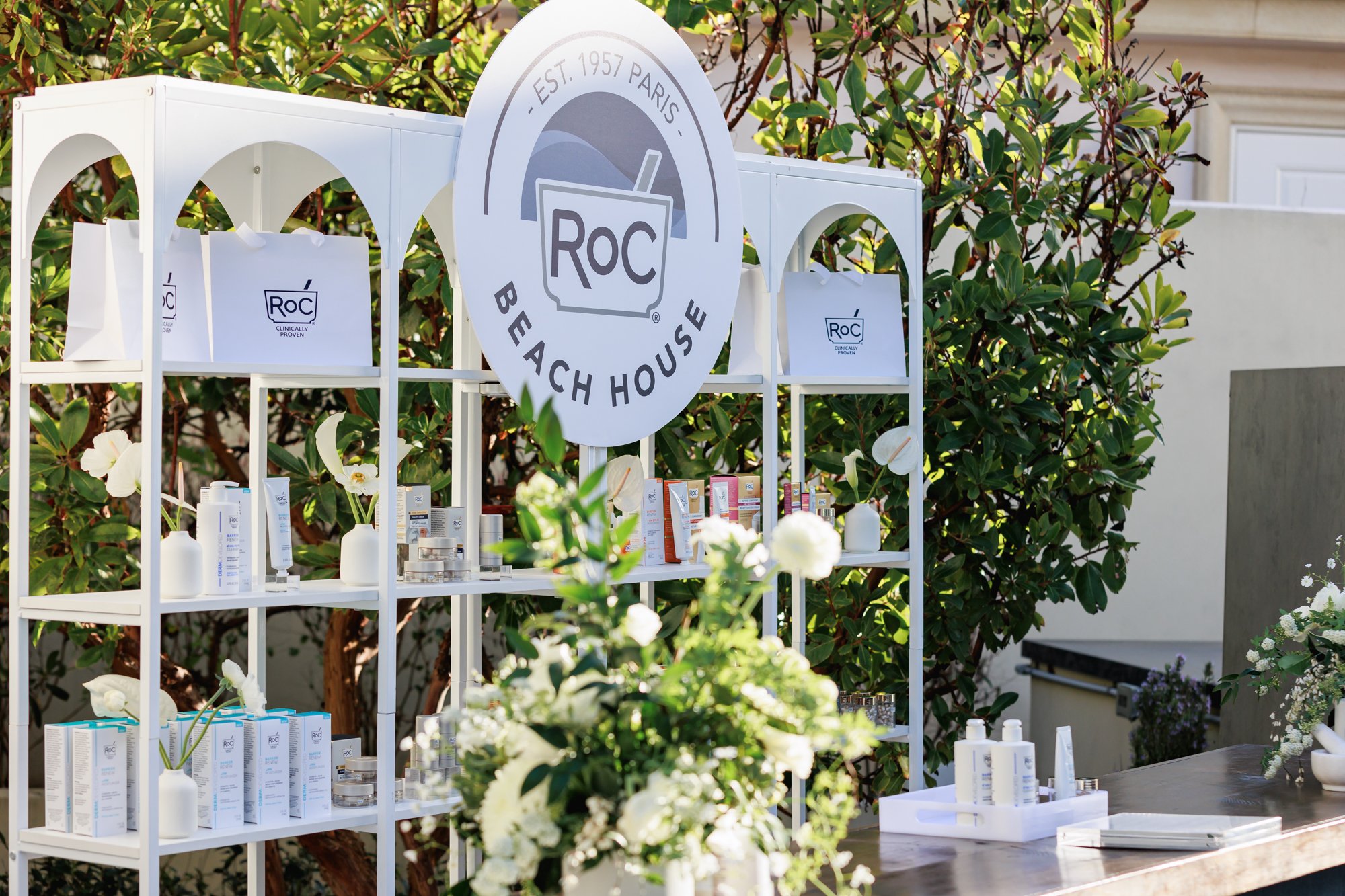 Outdoor display stand with Rox skincare products, surrounded by greenery and white flowers, featuring a large Rox Beach House sign with established date and location.