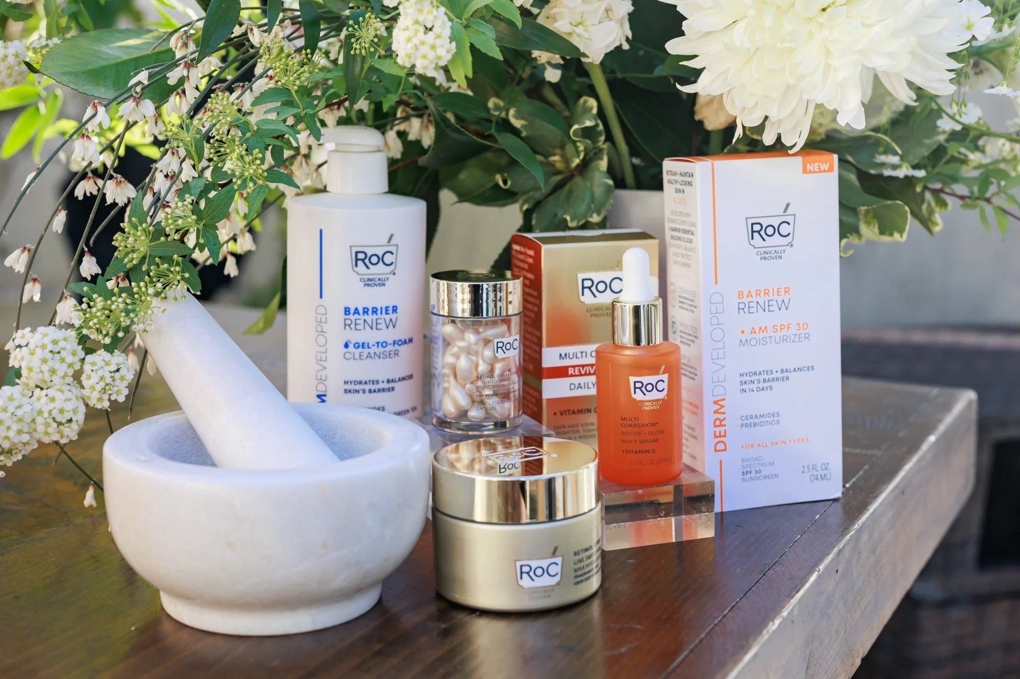 Beauty skincare products, including bottles and boxes, are arranged on a wooden surface amidst white flowers and green foliage.