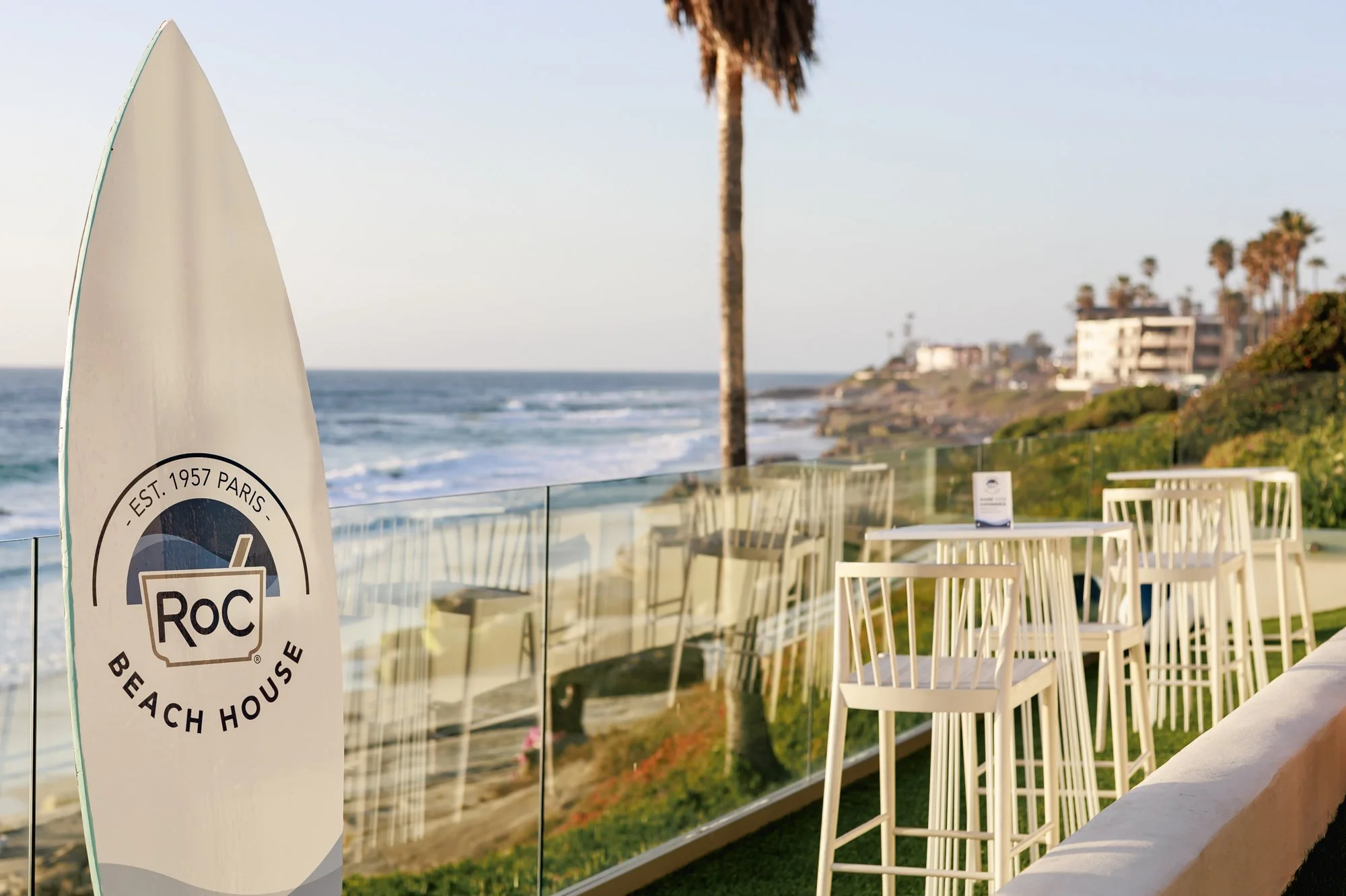 Beach house patio with white chairs, glass railing, palm trees, and ocean view during sunset, with a RoC beach house flag.